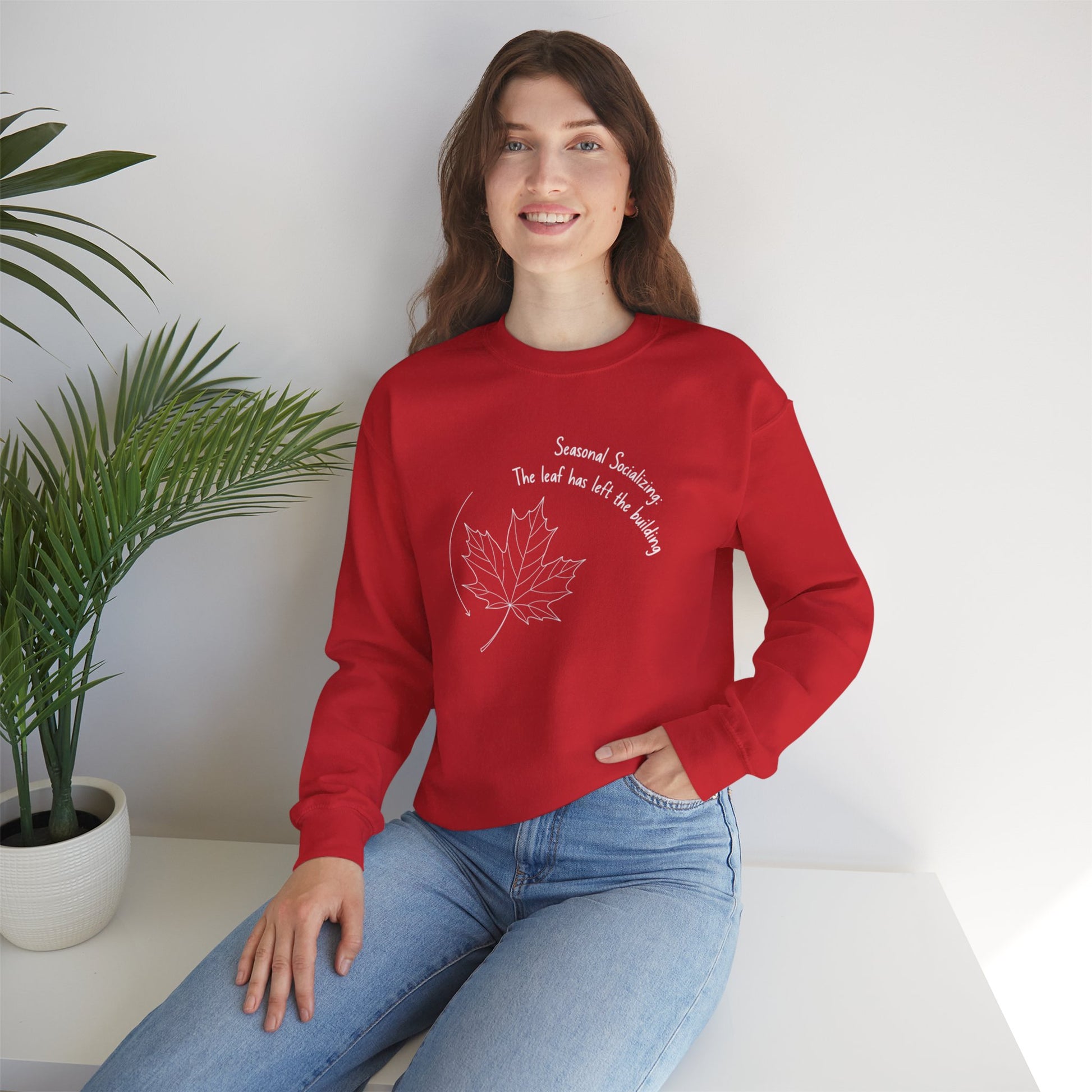 Person wearing a red sweatshirt with a leaf design and text 'Seasonal Socializing, The leaf has left the building', sitting on a white surface with a plant in the background.