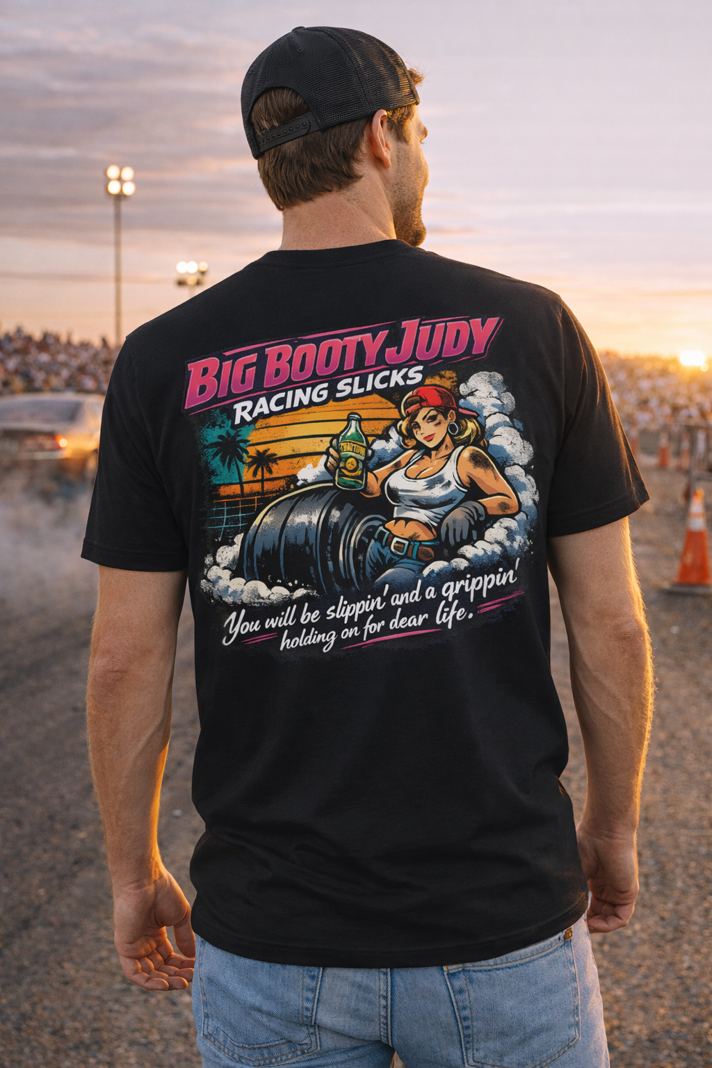 Man wearing a black t-shirt with a colorful graphic design and 'Big Booty Judy Racing Slicks You will be slippin' and a grippin' holding on for dear life' text, standing at a drag race