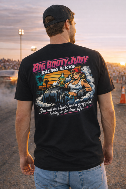 Man wearing a black t-shirt with a colorful graphic design and 'Big Booty Judy Racing Slicks You will be slippin' and a grippin' holding on for dear life' text, standing at a drag race