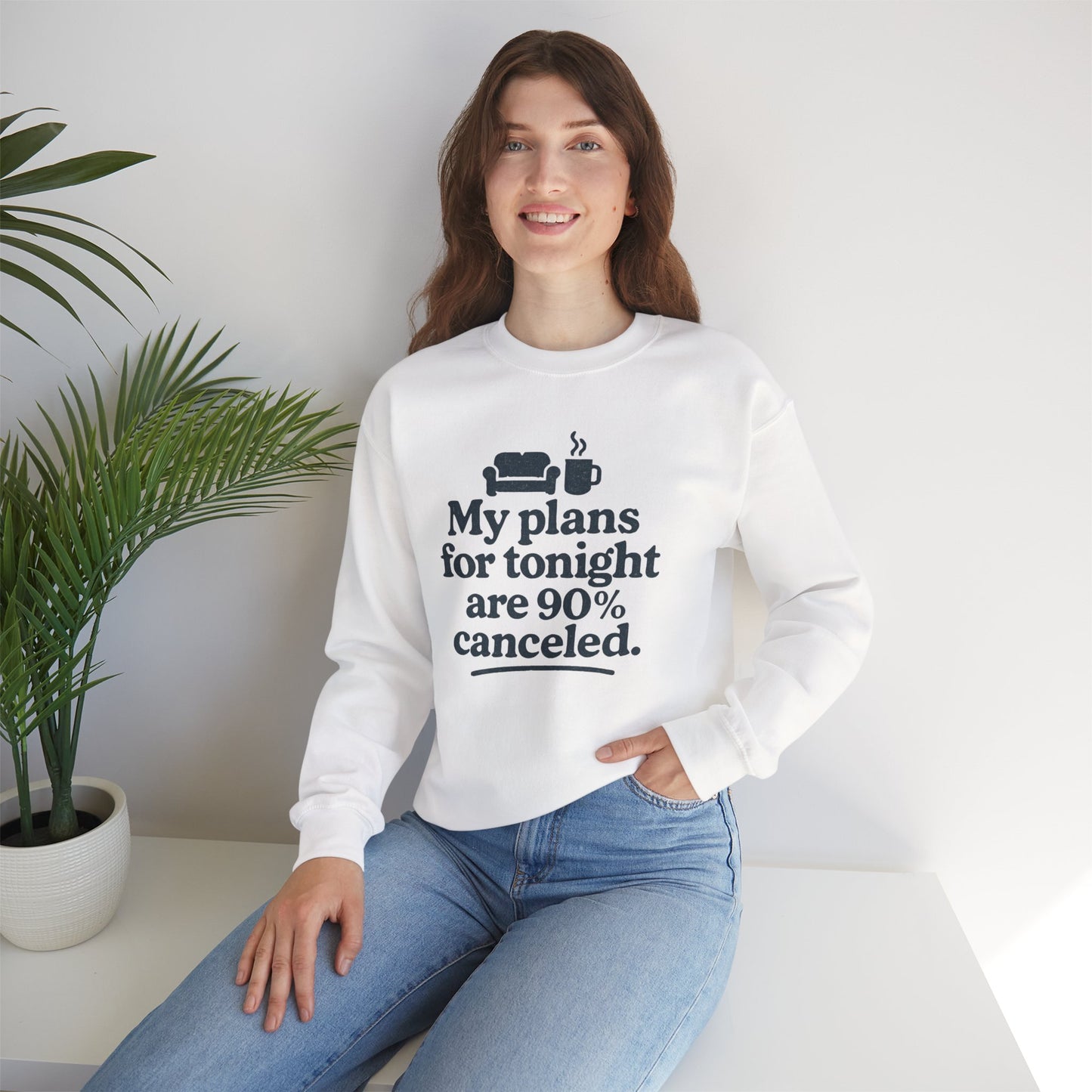 Woman wearing a white sweatshirt with text 'My plans for tonight are 90% canceled' sitting on a white surface with a plant in the background