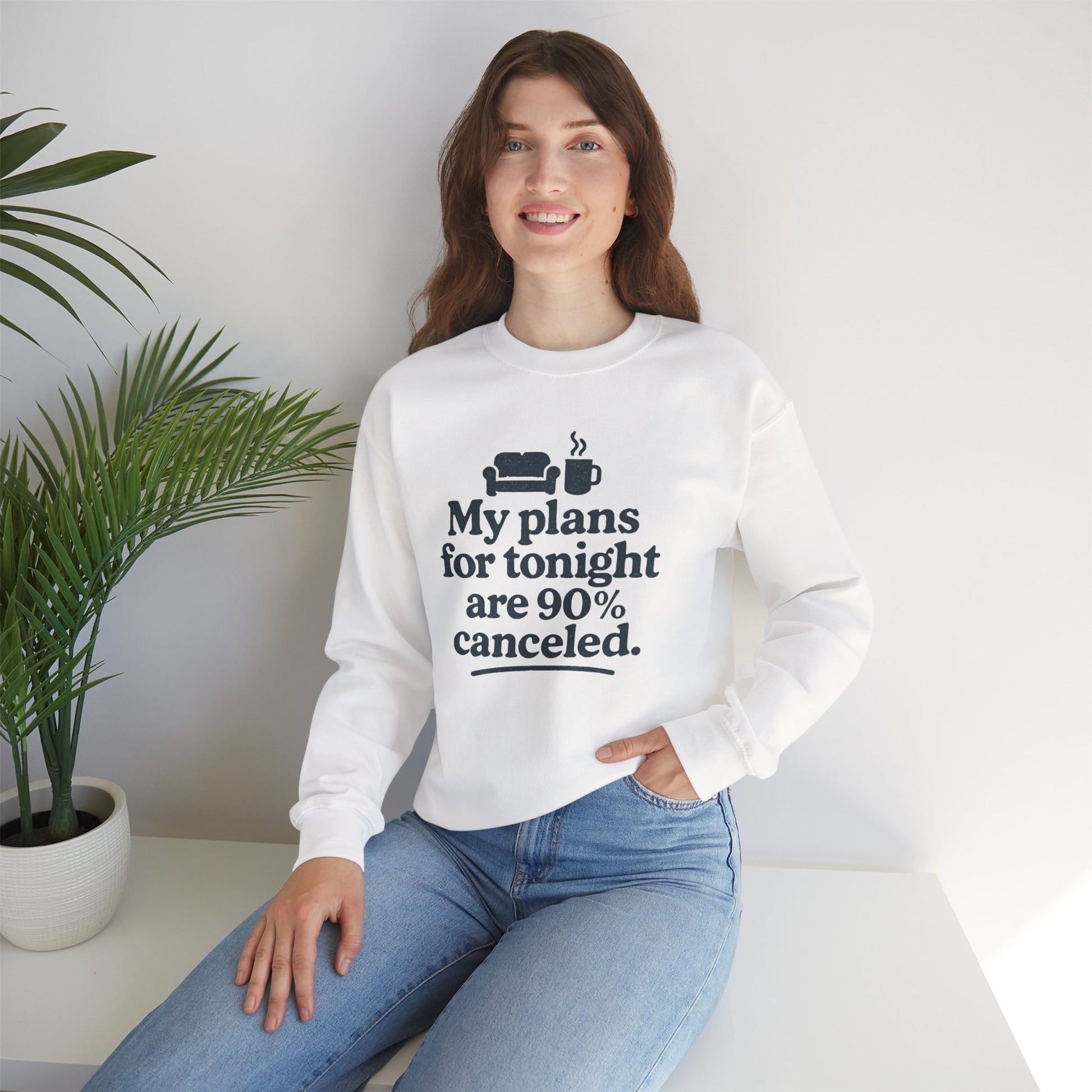 Woman wearing a white sweatshirt with text 'My plans for tonight are 90% canceled' sitting on a white surface with a plant in the background
