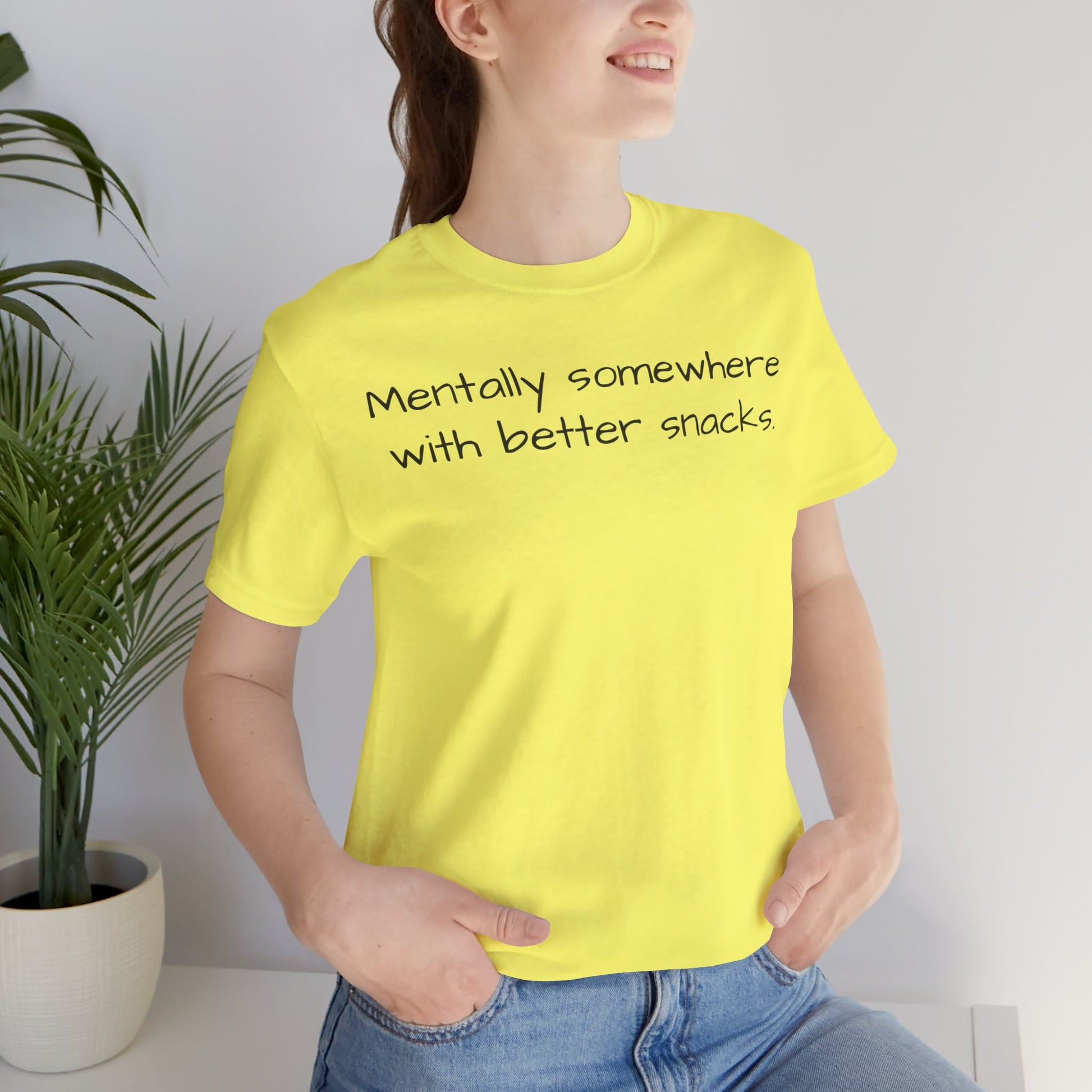 Person wearing a yellow t-shirt with text 'Mentally somewhere with better snacks', standing indoors next to a plant.