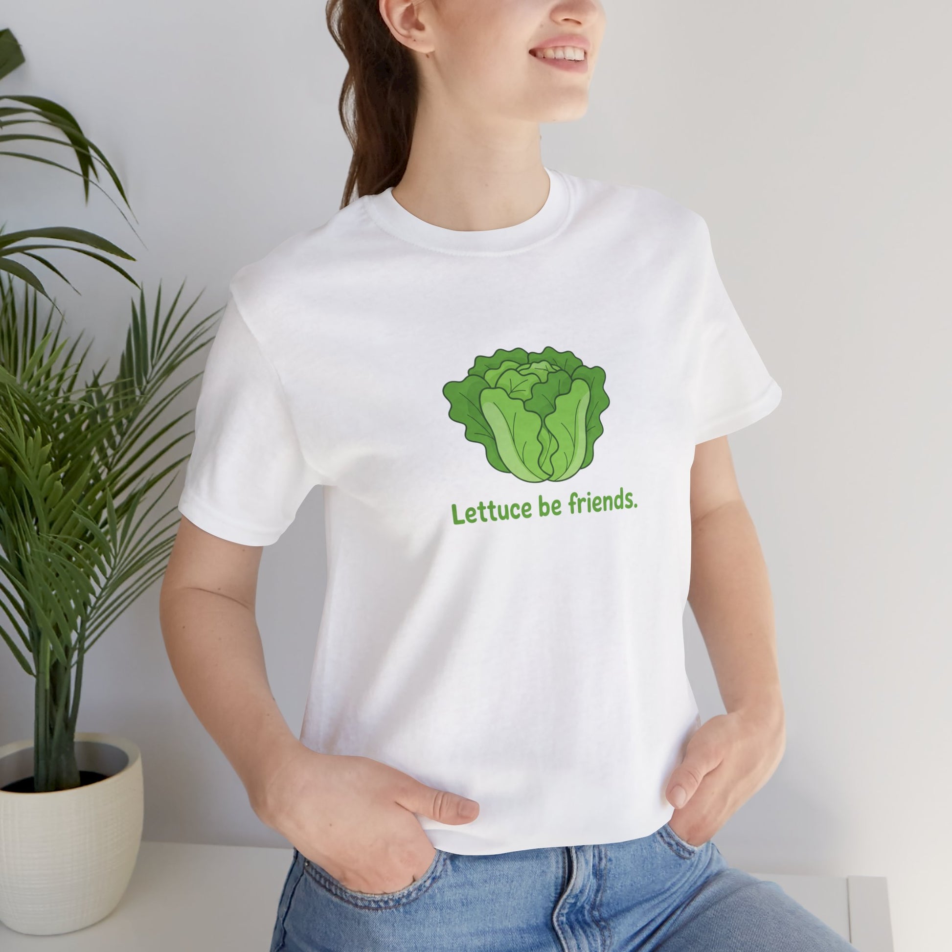 Person wearing a white t-shirt with a green lettuce graphic and text 'Lettuce be friends', standing indoors next to a plant.