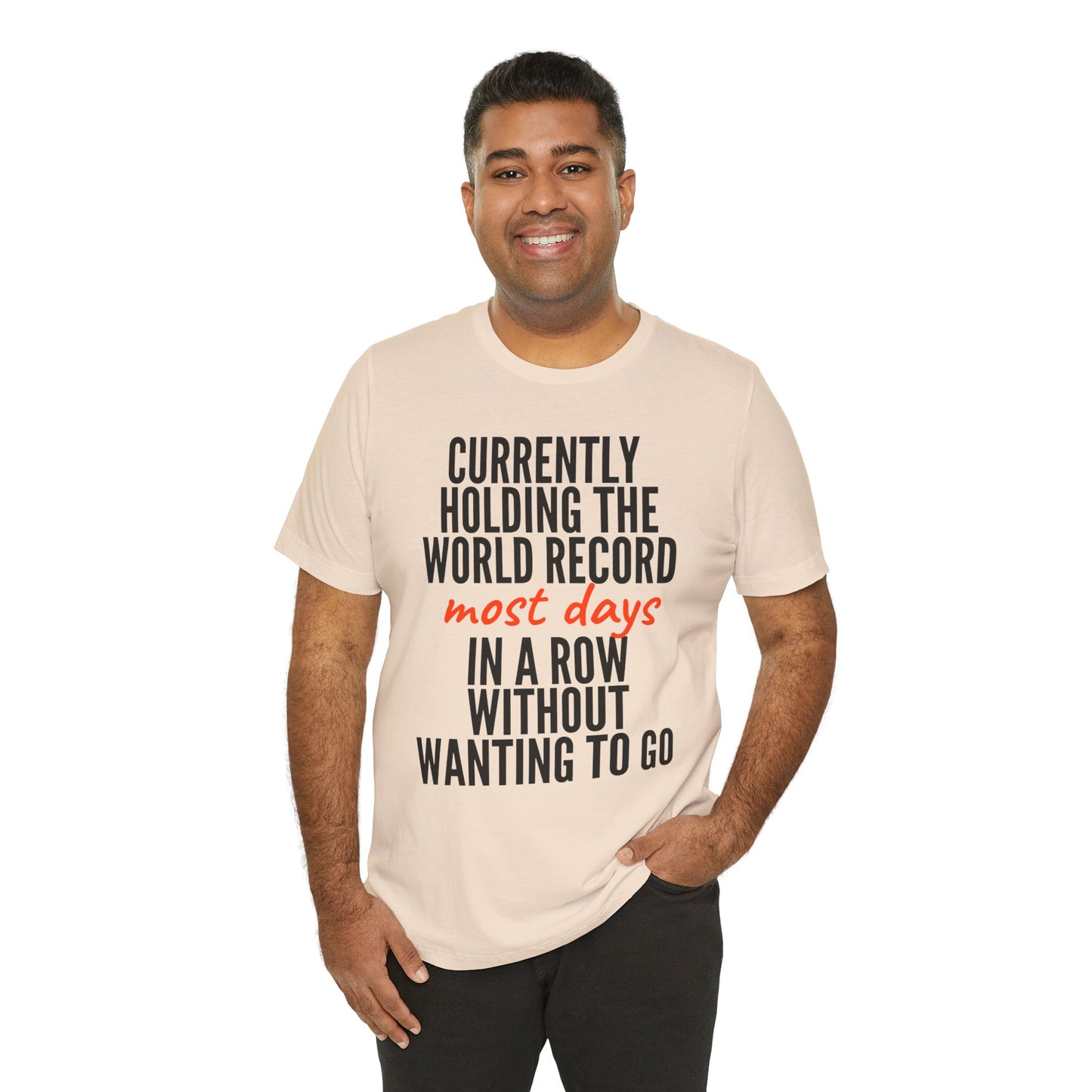 Man wearing a beige t-shirt with humorous text 'Currently holding the world record most days in a row without wanting to go' design on a white background