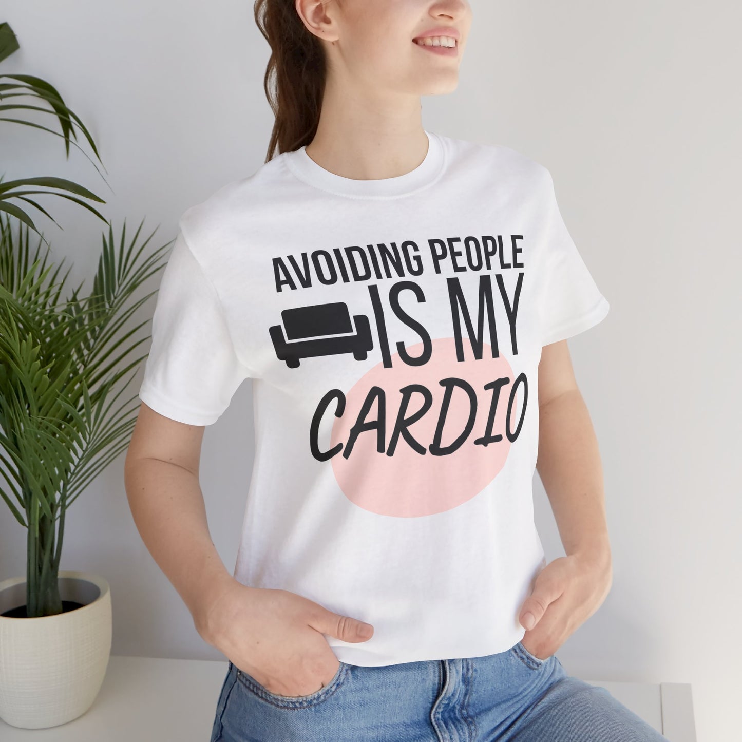 Person wearing a white t-shirt with humorous text 'Avoiding People Is My Cardio' design, standing indoors next to a plant.