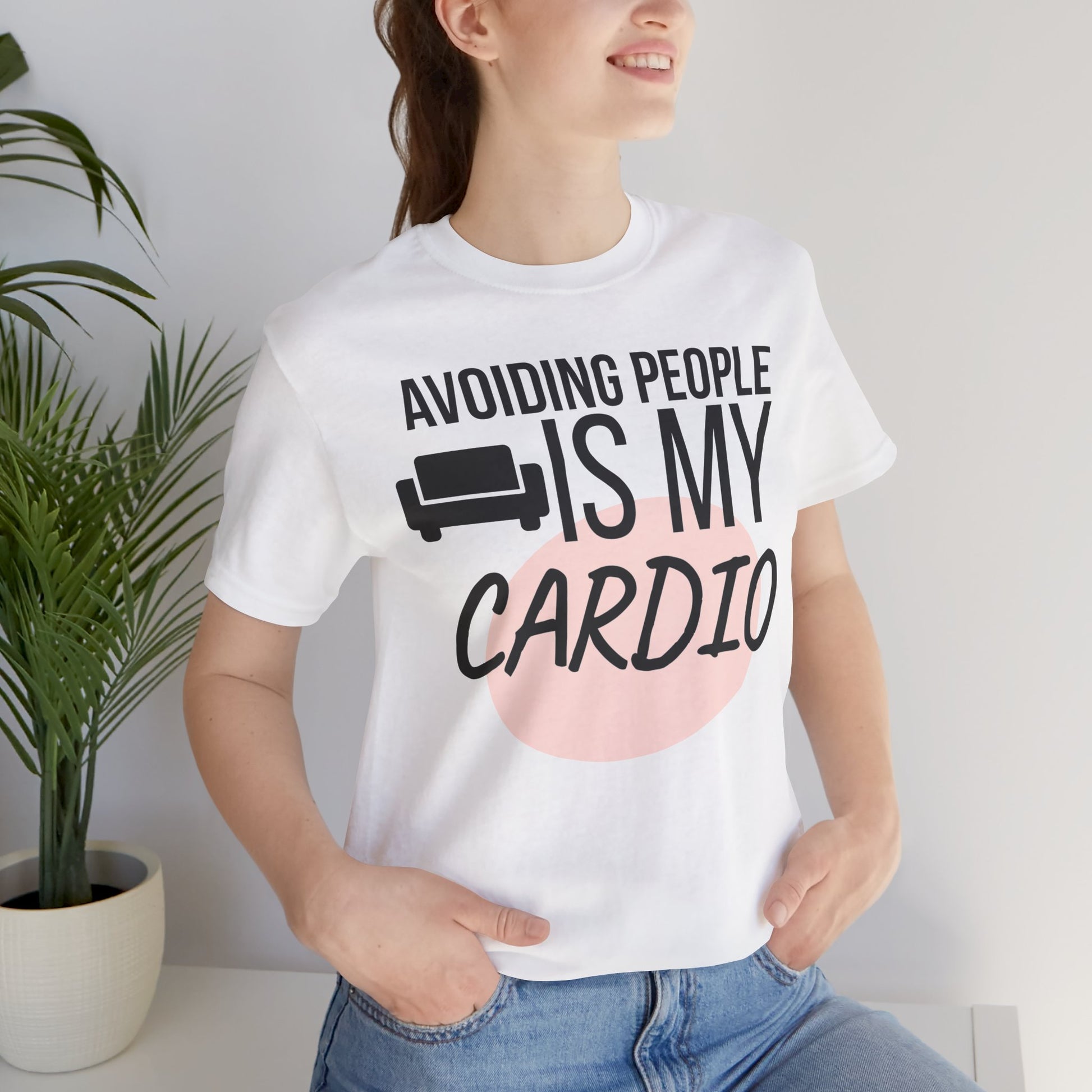 Person wearing a white t-shirt with humorous text 'Avoiding People Is My Cardio' design, standing indoors next to a plant.