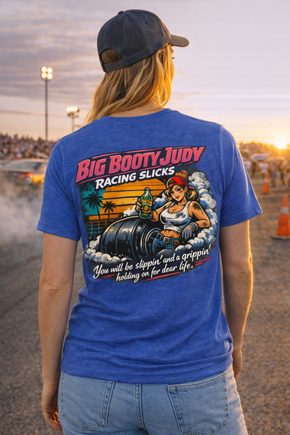 Woman wearing a heather true royal t-shirt with a colorful graphic design and 'Big Booty Judy Racing Slicks You will be slippin' and a grippin' holding on for dear life' text on the back, standing at a drag race