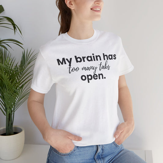Person wearing a white t-shirt with text 'My brain has too many tabs open.', standing indoors next to a plant.