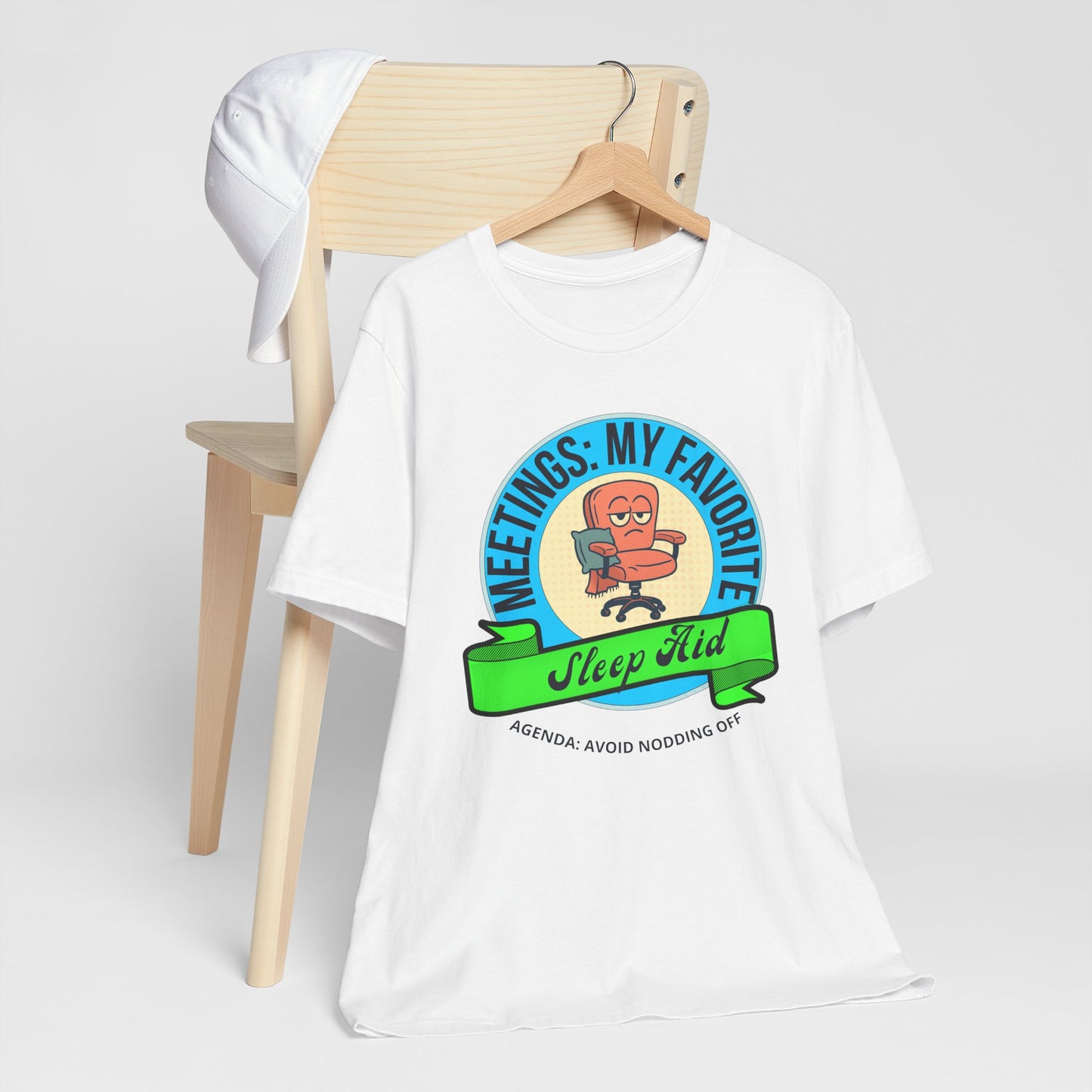 White t-shirt with a graphic design and text 'Meetings: My favorite sleep aid. Agenda: Avoid nodding off' on a wooden chair against a white background