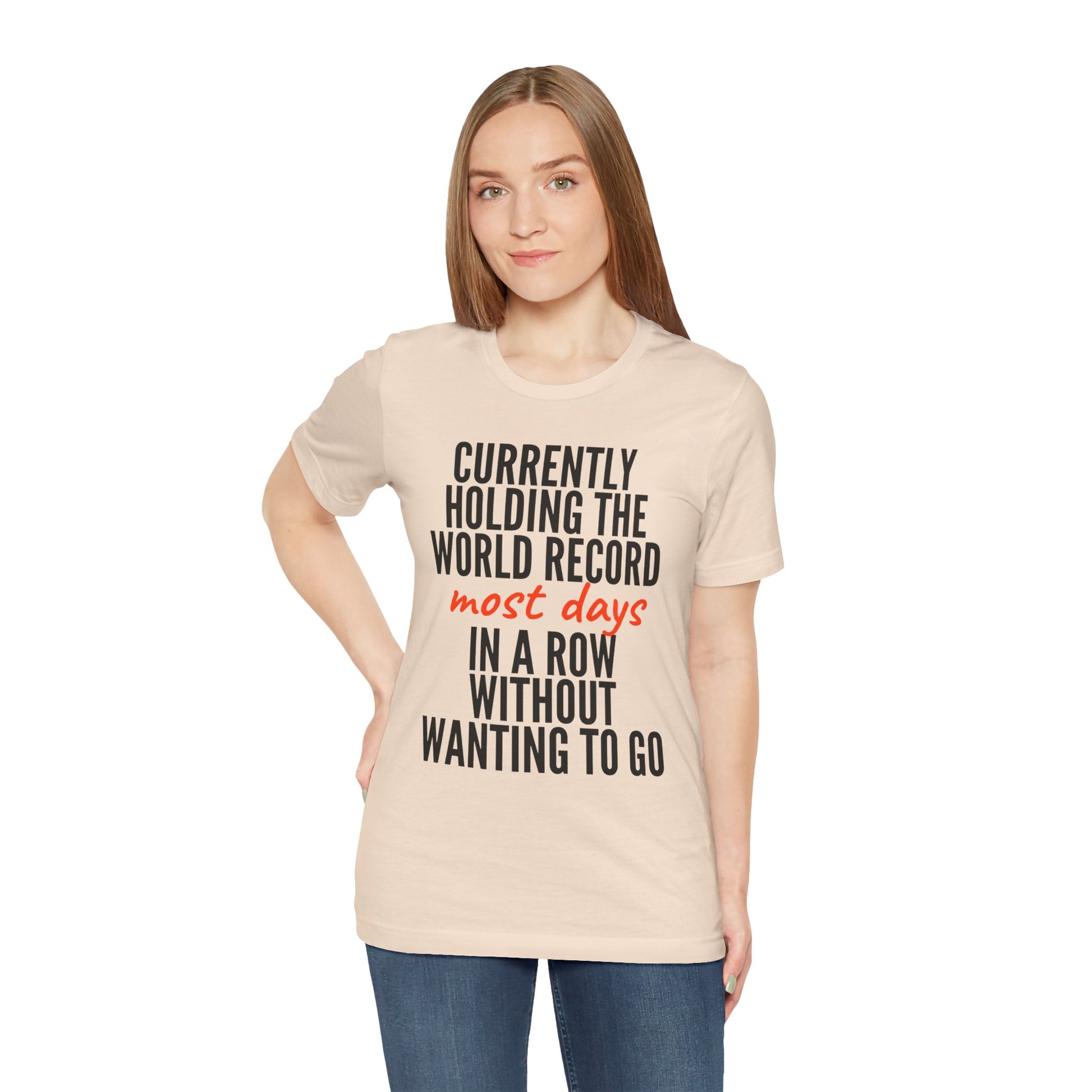 Woman wearing a beige t-shirt with humorous text 'Currently holding the world record most days in a row without wanting to go' design on a white background
