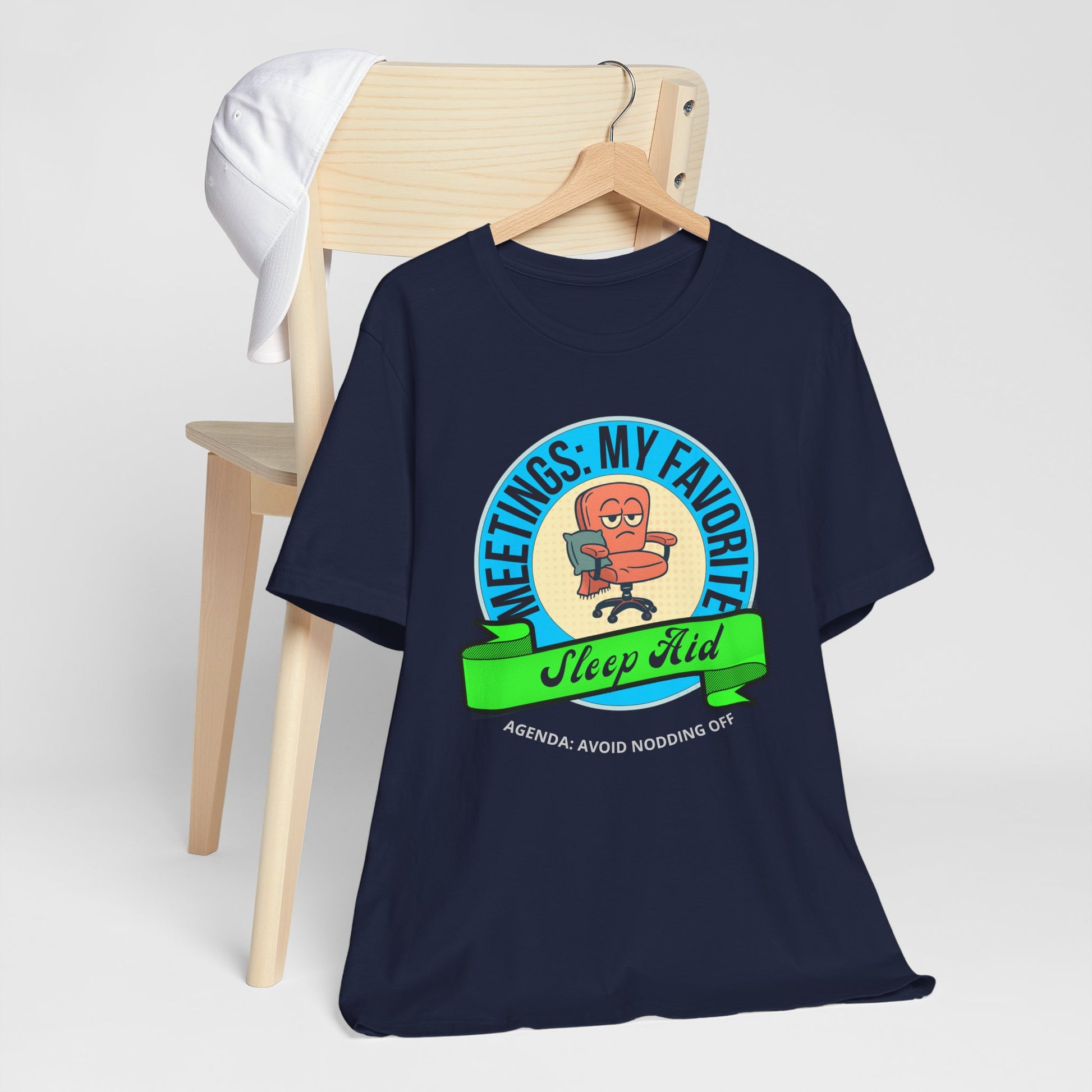 Navy blue t-shirt with graphic design and text 'Meetings: My favorite sleep aid. Agenda: Avoid nodding off' on a wooden chair against a white background