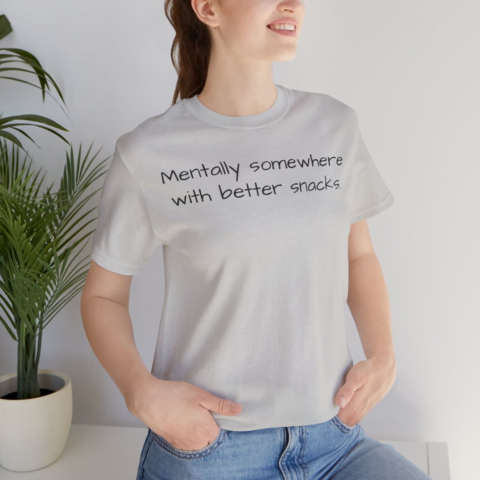 Person wearing a gray t-shirt with text 'Mentally somewhere with better snacks', standing indoors next to a plant.