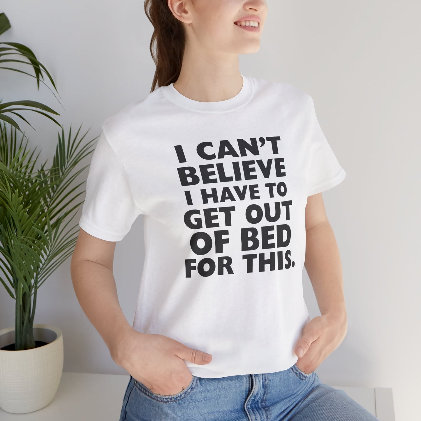 Person wearing a white t-shirt with black text 'I can't believe I have to get out of bed for this' on a white background