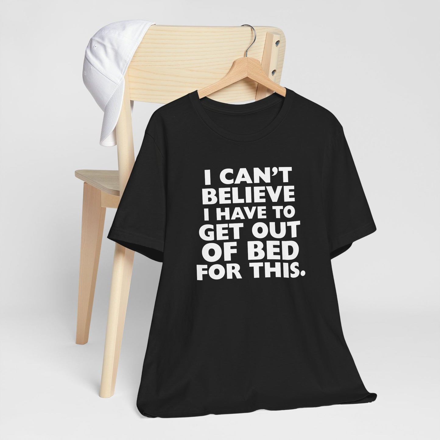 Black t-shirt with humorous text 'I can't believe I have to get out of bed for this' on a hanger against a white background
