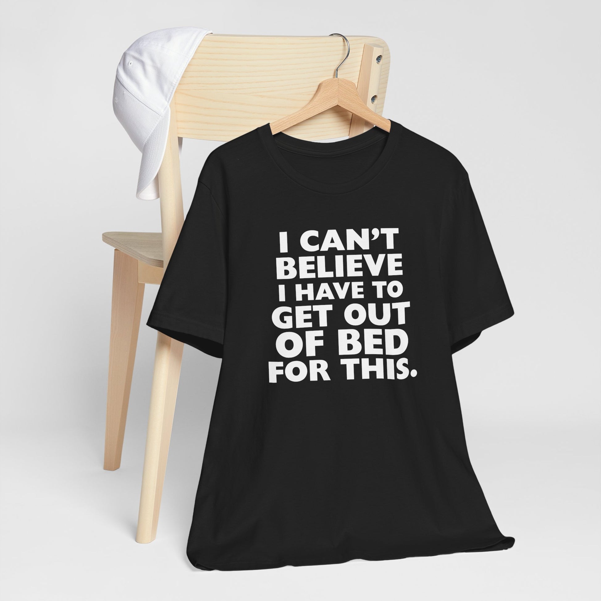Black t-shirt with humorous text 'I can't believe I have to get out of bed for this' on a hanger against a white background