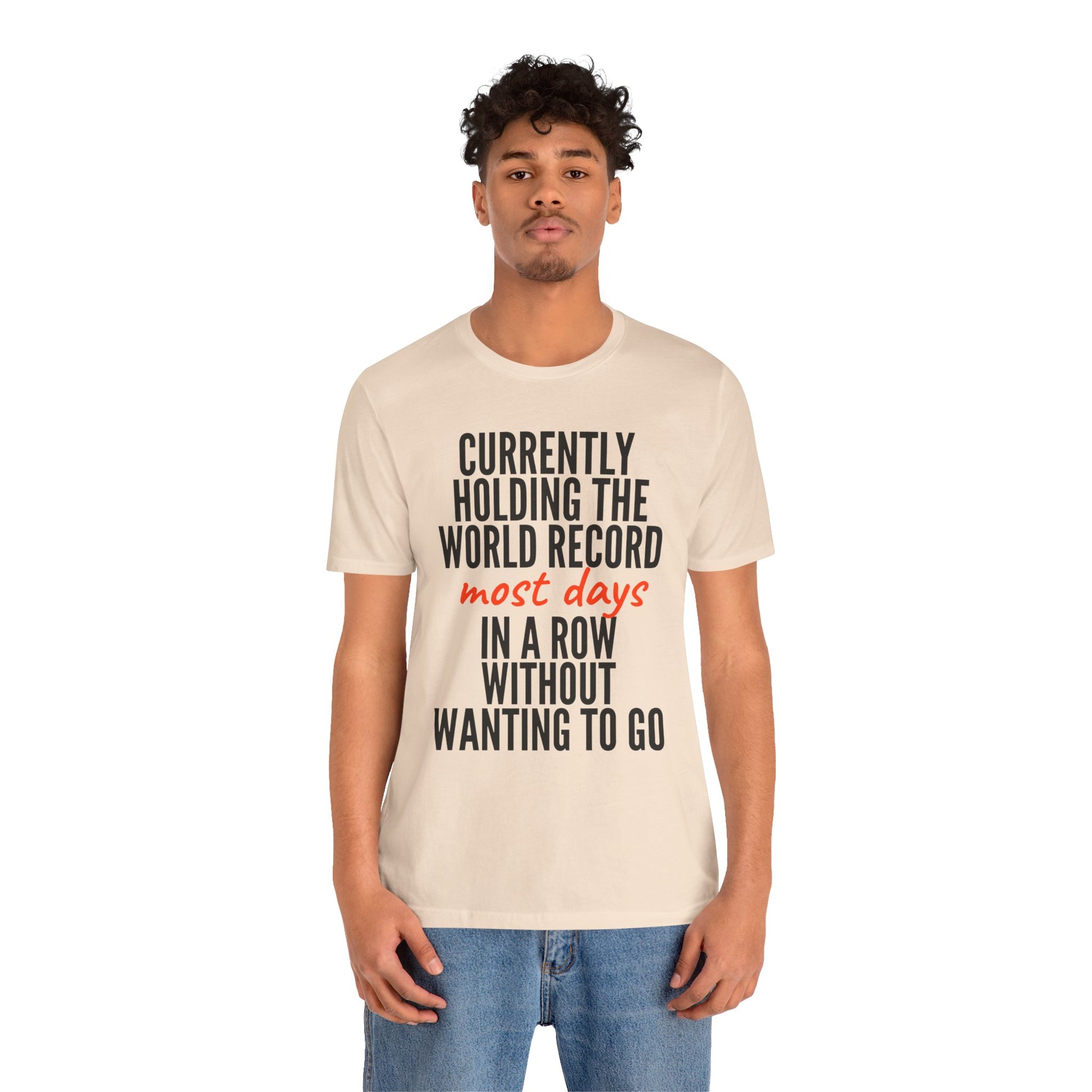 Man wearing a beige t-shirt with humorous text 'Currently holding the world record most days in a row without wanting to go' design on a white background