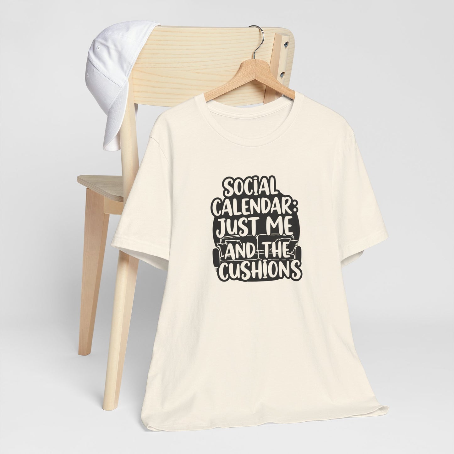 Beige t-shirt with text 'Social Calendar: Just Me and the Cushions' on a wooden chair.