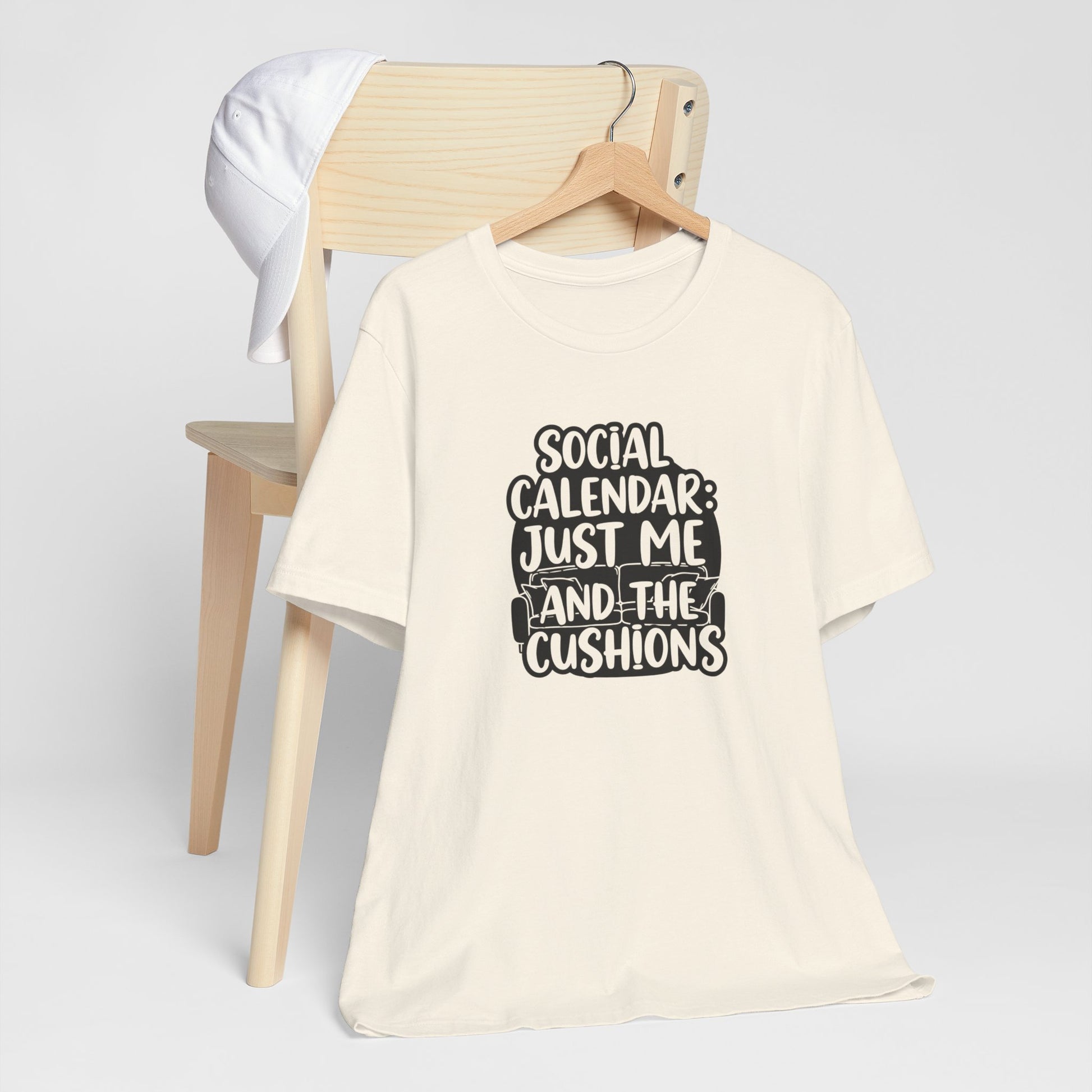Beige t-shirt with text 'Social Calendar: Just Me and the Cushions' on a wooden chair.