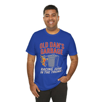 Person wearing a blue graphic T‑shirt featuring the bold red and white text “Old Dan’s Garbage” above a flaming garbage can illustration, with the tagline “Racing Junk in the Trunk” below. Funny drag racing parody shirt with retro racing flames, checkered flag background, and humor‑driven hot rod style, perfect for car enthusiasts and gearhead apparel fans.