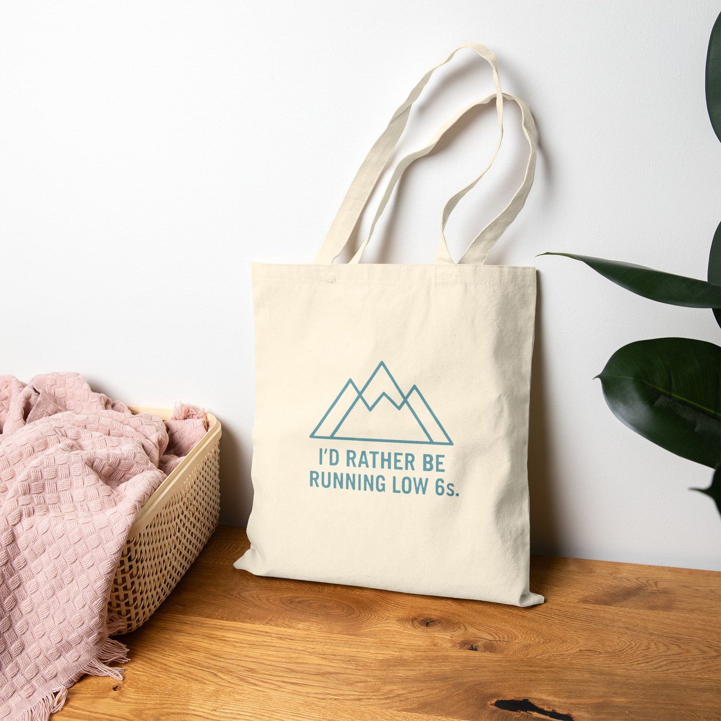 Tote bag with mountain design and text 'I'd rather be running low 6s' on a wooden surface with a white wall and plant in the background