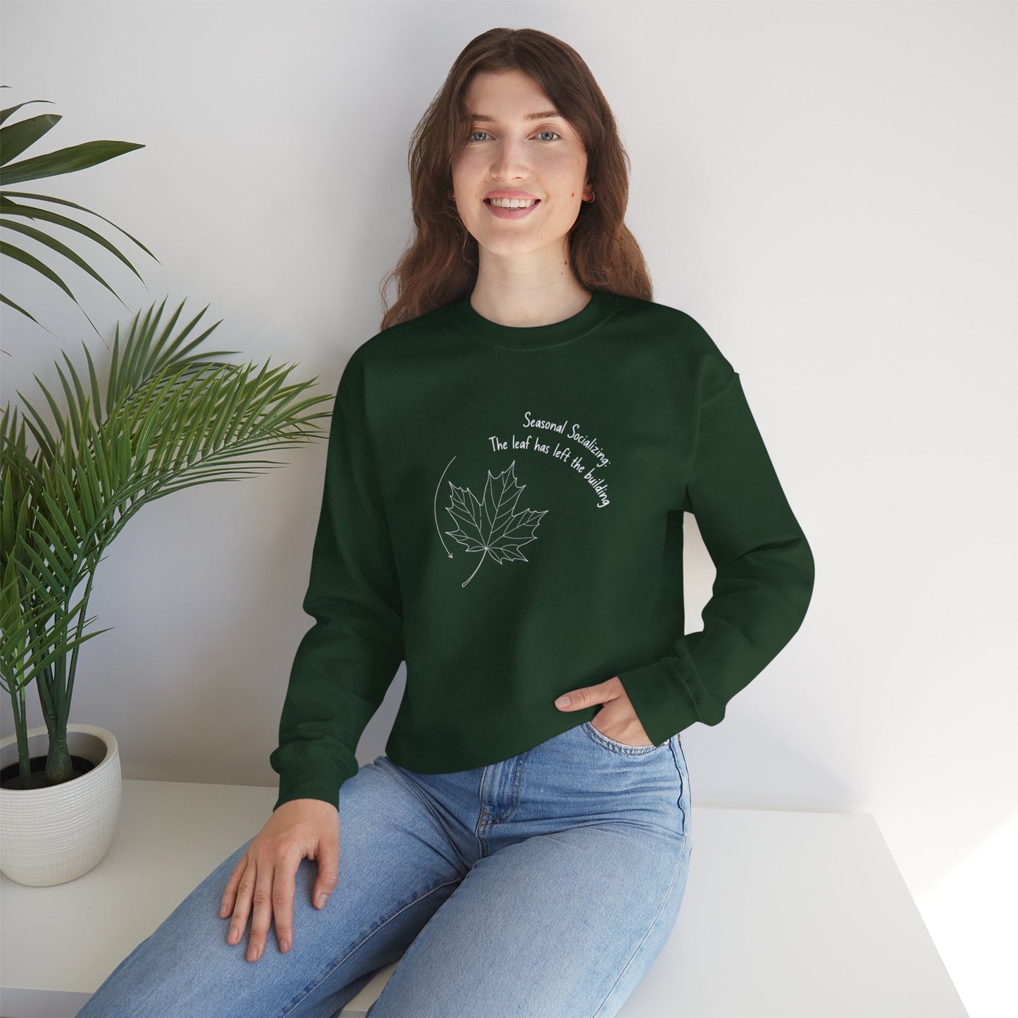 Person wearing a green sweatshirt with a leaf design and text 'Seasonal Socializing, The leaf has left the building', sitting on a white surface with a plant in the background.