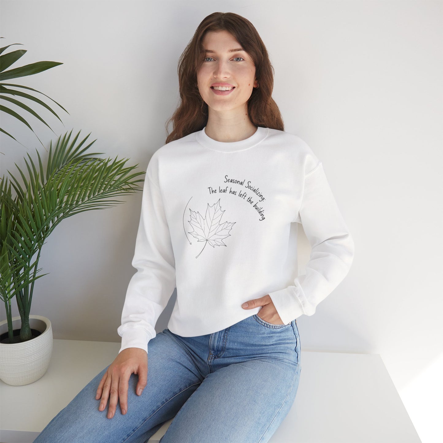 Woman wearing a white sweatshirt with text 'Seasonal Socializing, The leaf has left the building'and a design, sitting in a room with a plant.