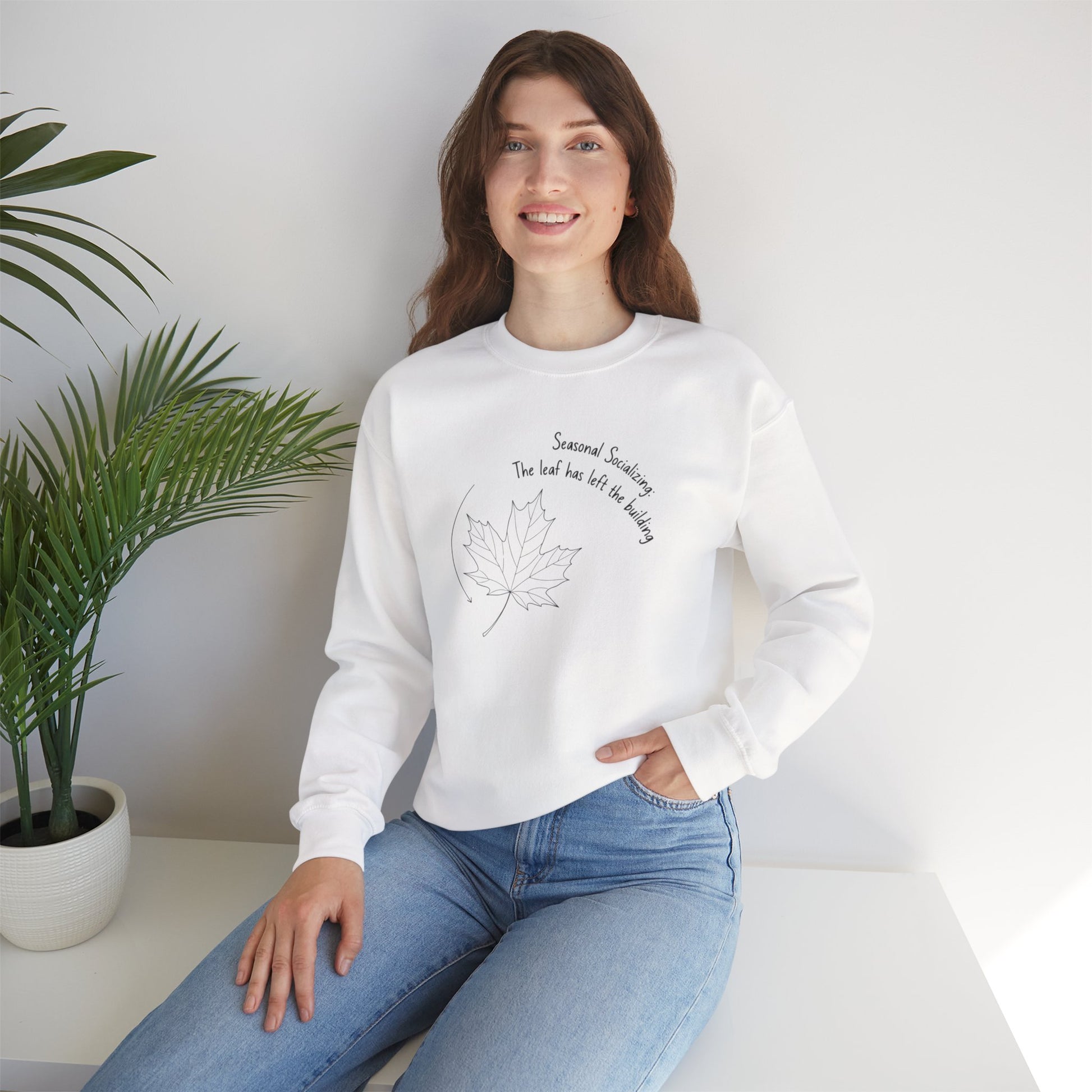 Woman wearing a white sweatshirt with text 'Seasonal Socializing, The leaf has left the building'and a design, sitting in a room with a plant.