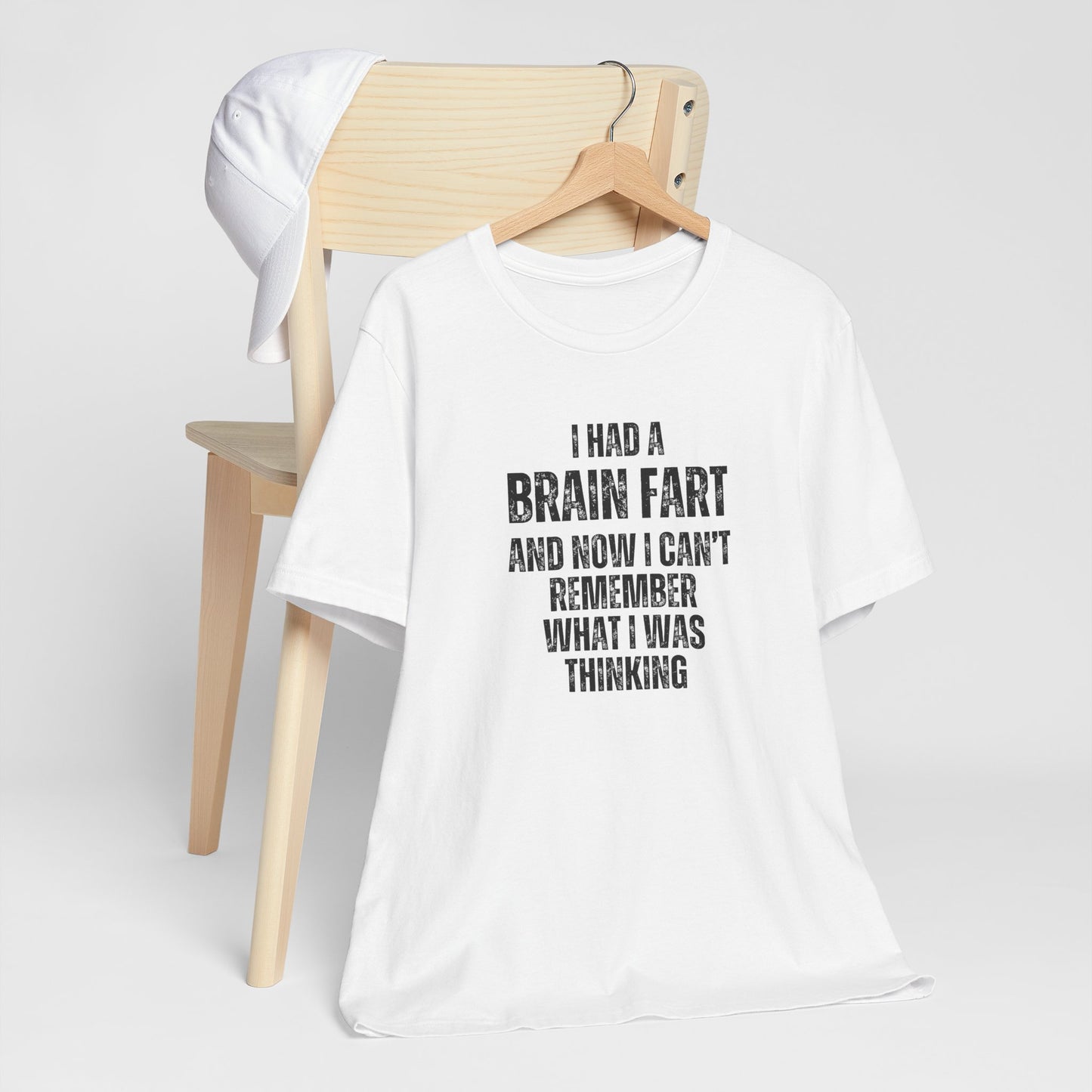 White t-shirt with humorous text 'I had a brain fart and now I can't remember what I was thinking' on a wooden chair against a white background