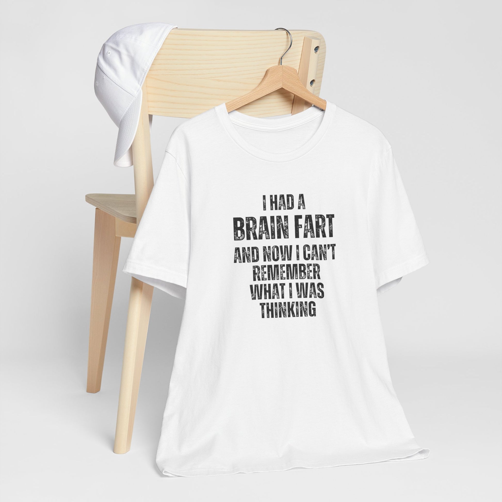 White t-shirt with humorous text 'I had a brain fart and now I can't remember what I was thinking' on a wooden chair against a white background