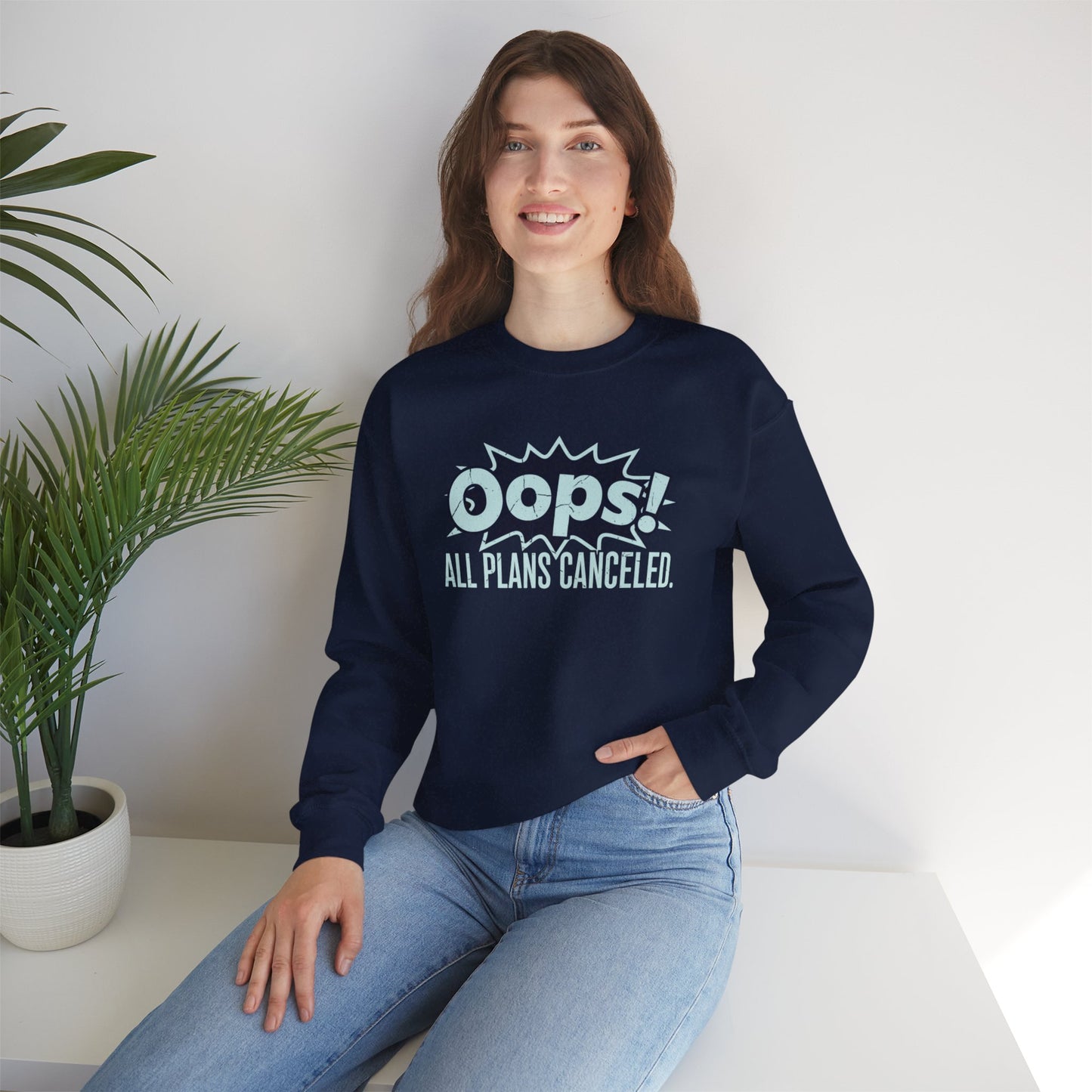 Person wearing a navy blue sweatshirt with 'Oops! All plans canceled' text, sitting on a white surface with a plant in the background.