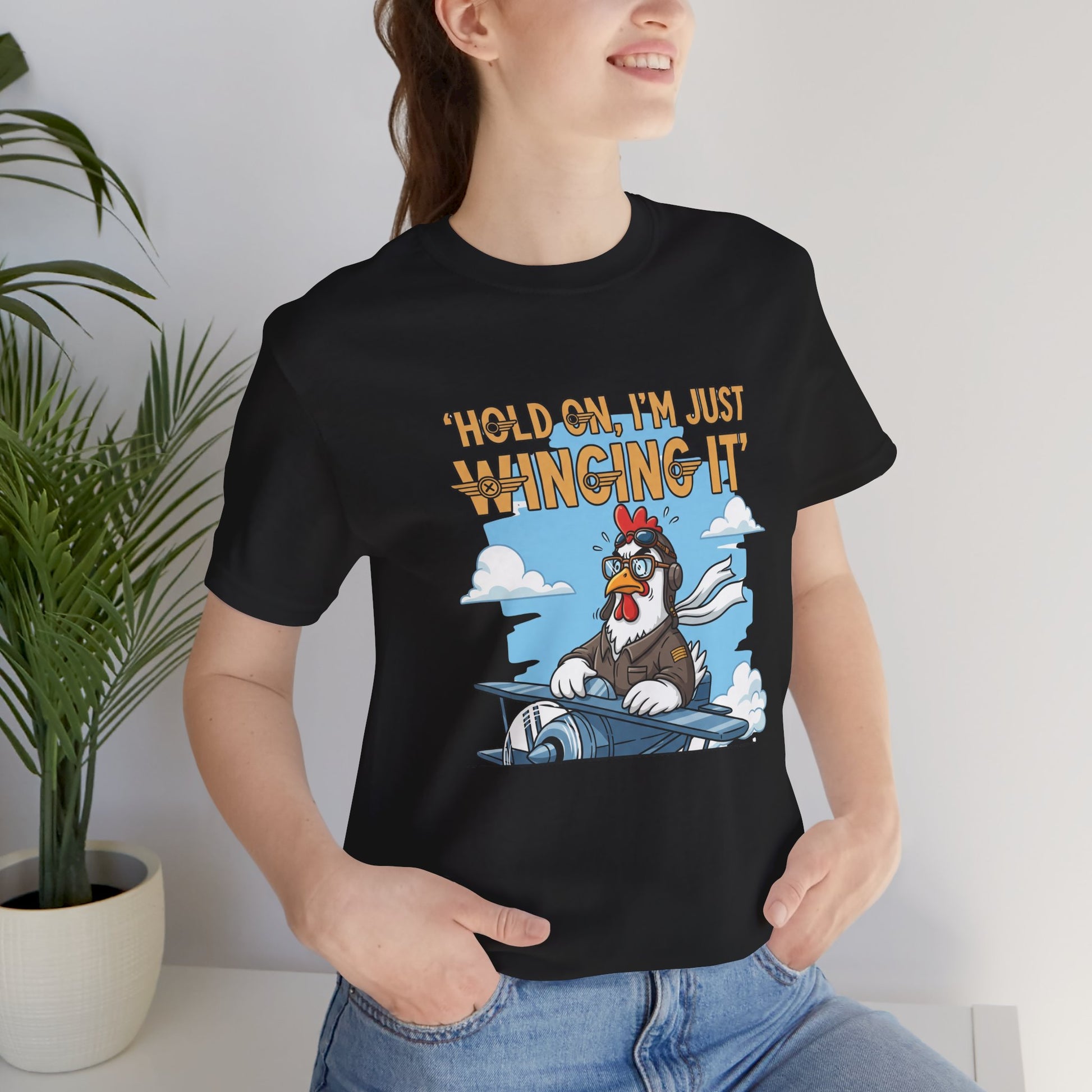 Person wearing a black t-shirt with a cartoon chicken graphic and text 'Hold on, I'm just winging it', standing indoors.