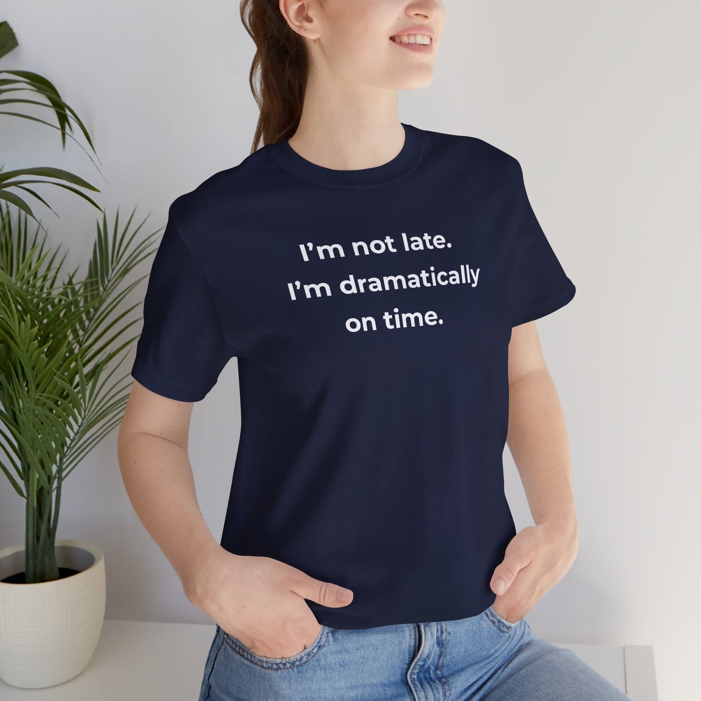 Person wearing a navy blue t-shirt with humorous text 'I'm not late. I'm dramatically on time', standing indoors next to a plant.