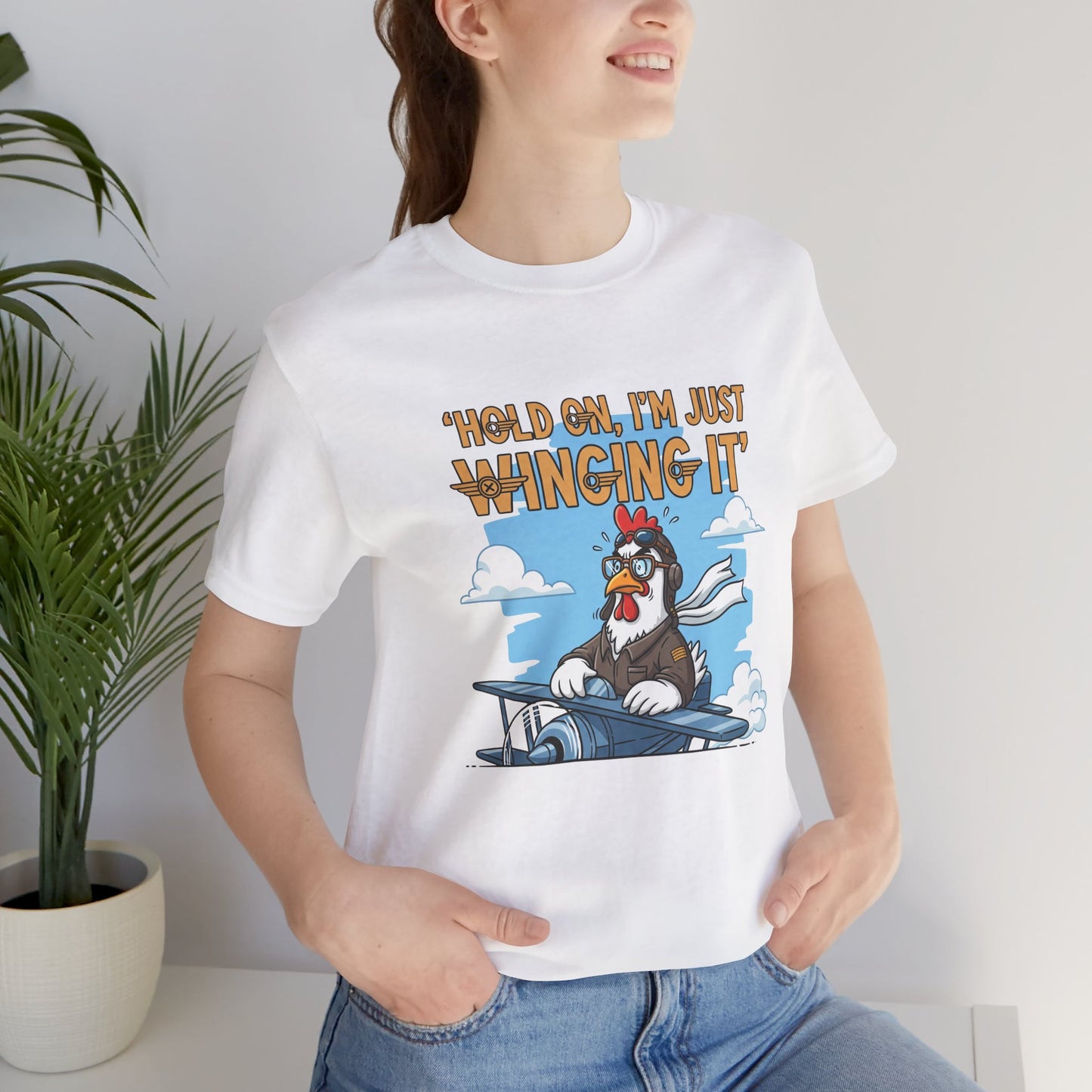 Person wearing a white t-shirt with a cartoon chicken on a plane graphic and text 'Hold on, I'm just winging it'.