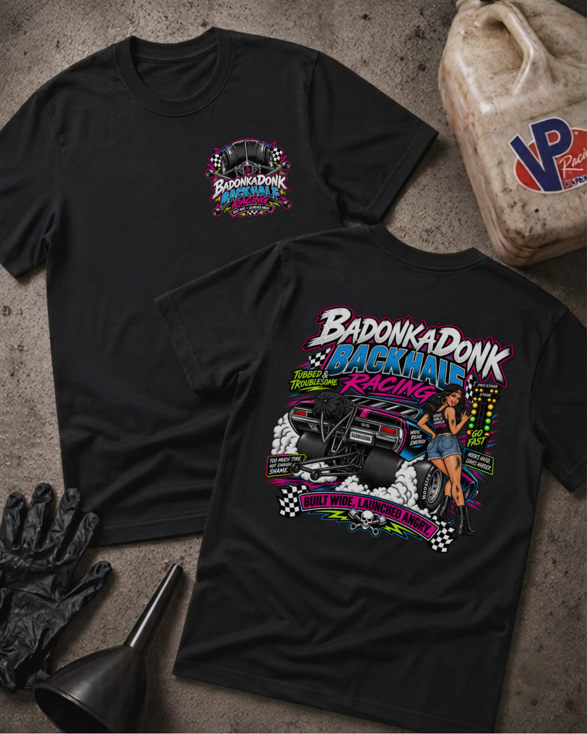 Two black graphic t-shirts displayed flat on a textured gray surface, surrounded by racing-themed props including a jerrycan, black gloves, and a funnel. The front shirt features a small 'Badonkadonk Racing' chest logo, while the back shirt displays a large, colorful graphic of a race car and a pin-up girl with the text 'Badonkadonk Backhalf Racing'.