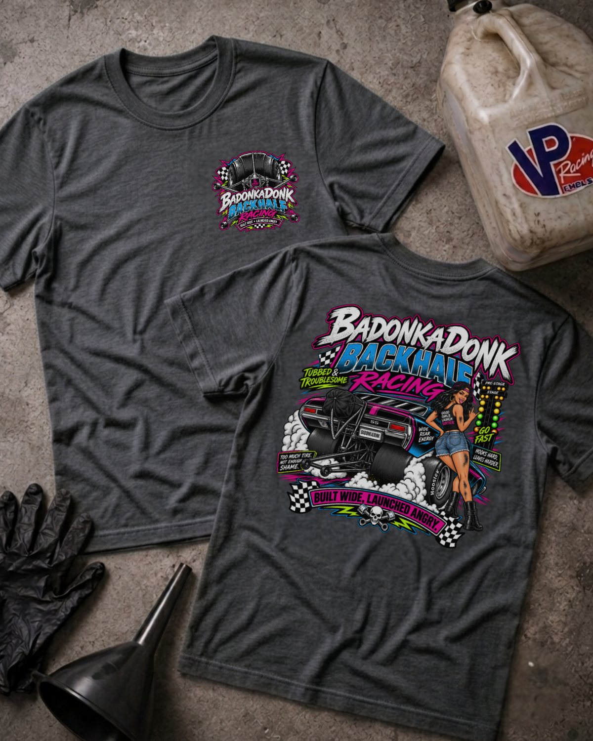Two dark grey heather graphic t-shirts displayed flat on a textured gray surface, surrounded by racing-themed props including a jerrycan, black gloves, and a funnel. The front shirt features a small 'Badonkadonk Racing' chest logo, while the back shirt displays a large, colorful graphic of a race car and a pin-up girl with the text 'Badonkadonk Backhalf Racing'.