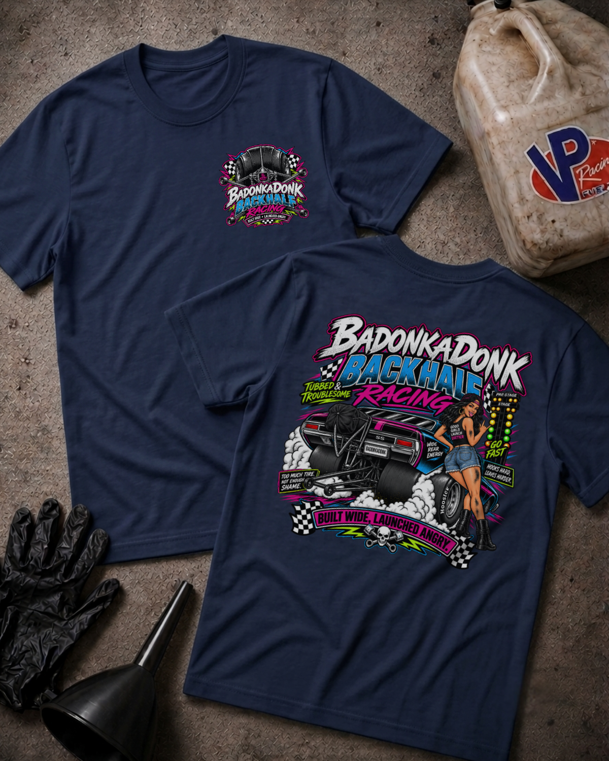 Two navy blue graphic t-shirts displayed flat on a textured gray surface, surrounded by racing-themed props including a jerrycan, black gloves, and a funnel. The front shirt features a small 'Badonkadonk Racing' chest logo, while the back shirt displays a large, colorful graphic of a race car and a pin-up girl with the text 'Badonkadonk Backhalf Racing'.