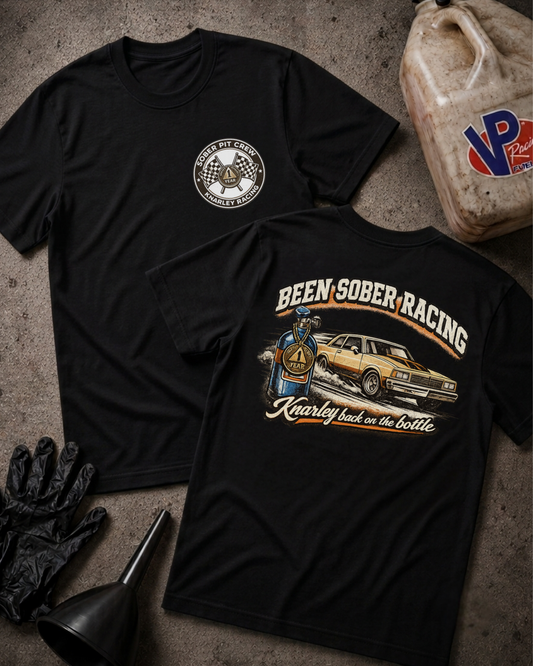 Two black graphic t-shirts displayed on a textured concrete surface with racing-themed props, including a gas can, gloves, funnel, and a race medallion. One shirt shows a circular chest logo, and the other shows a large back graphic reading 'Been Sober Racing' with a race car and driver illustration.