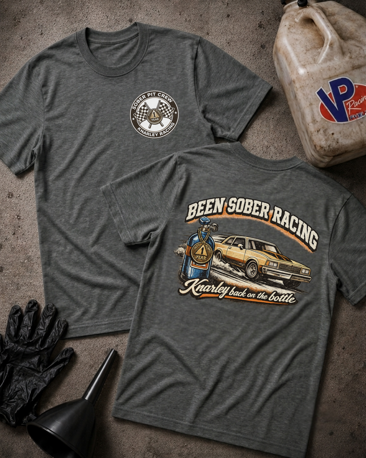 Two heather dark gray graphic t-shirts laid flat on a textured concrete-like surface, with gloves, funnel, and a plastic jug around them. The front shirt shows a small circular chest graphic reading 'SOBER PIT CREW KNARLEY RACING', and the back shirt shows a large graphic reading 'BEEN SOBER RACING' with a car and bottle illustration and the phrase 'Kearley back in the bottle'.