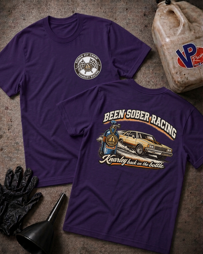 Two purple short-sleeve t-shirts laid flat on a textured concrete-like surface, showing the front and back designs. The front shirt has a small circular chest graphic, and the back shirt has a large graphic reading 'Been Sober Racing' with a car, bottle, and decorative text.