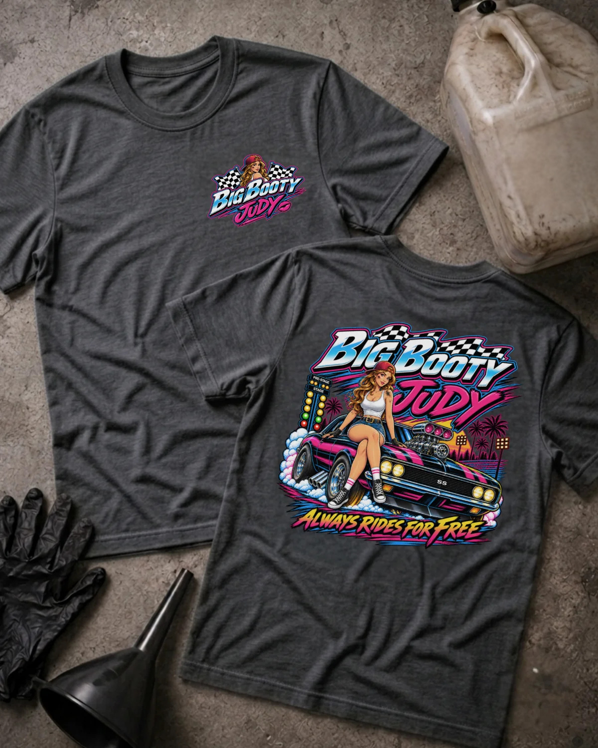 Two dark grey heather  graphic T-shirts laid flat on a textured gray surface, with a vintage gas can, black gloves, and a black funnel as styled props. The front shirt shows a small multicolor 'Big Booty Judy' chest graphic, and the back shirt shows a large colorful graphic of a woman standing on a pink car with the text 'Big Booty Judy' and 'Always Rides For Free'.