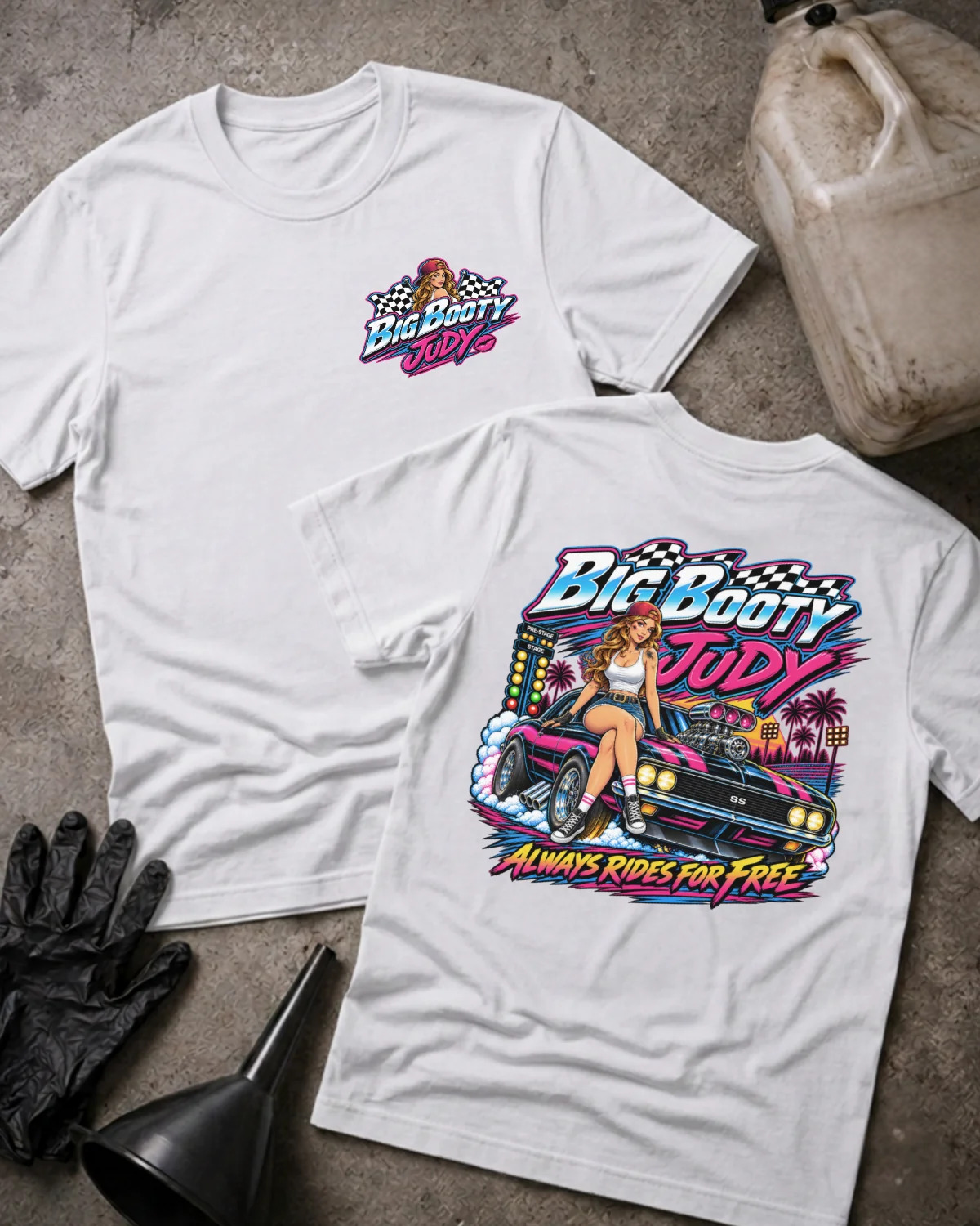 Two white  graphic T-shirts laid flat on a textured gray surface, with a vintage gas can, black gloves, and a black funnel as styled props. The front shirt shows a small multicolor 'Big Booty Judy' chest graphic, and the back shirt shows a large colorful graphic of a woman standing on a pink car with the text 'Big Booty Judy' and 'Always Rides For Free'.