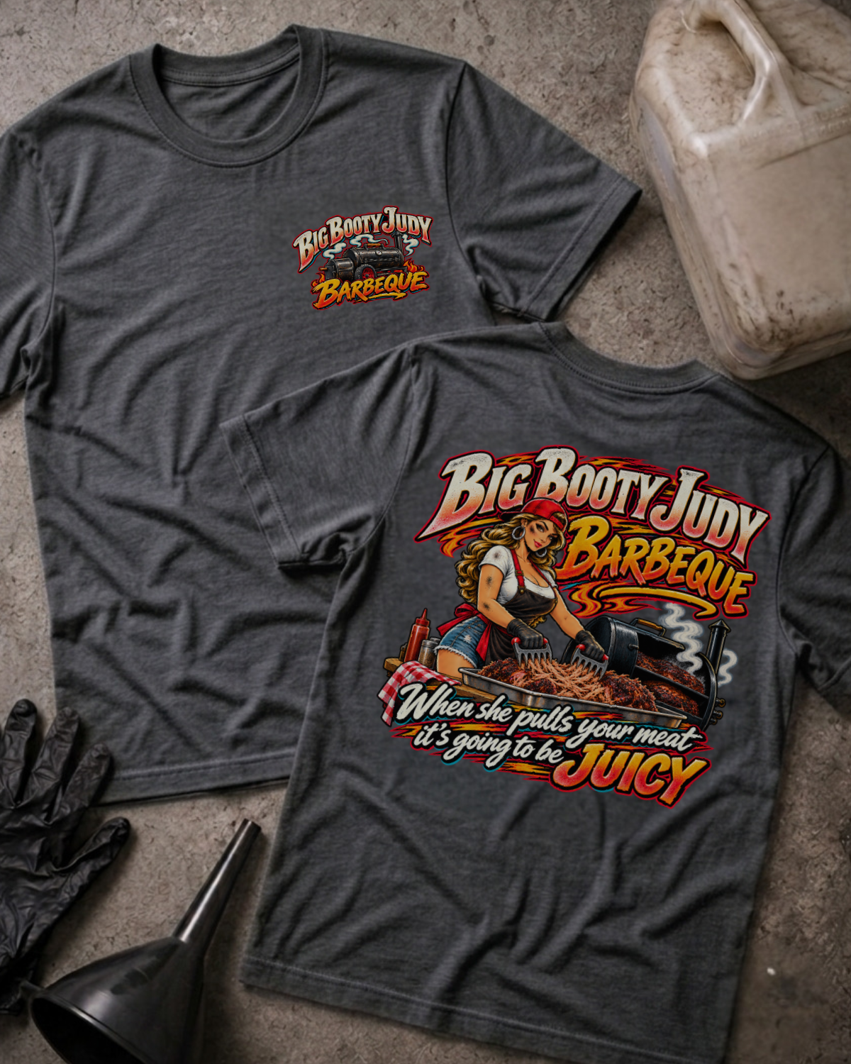 Big Booty Judy Barbeque Juicy T-shirt in dark grey heather, flat lay showing the front chest logo and the back graphic reading Big Booty Judy Barbeque, When She Pulls Your Meat It's Going to Be Juicy, styled with work gloves, a funnel, and a fluid jug.