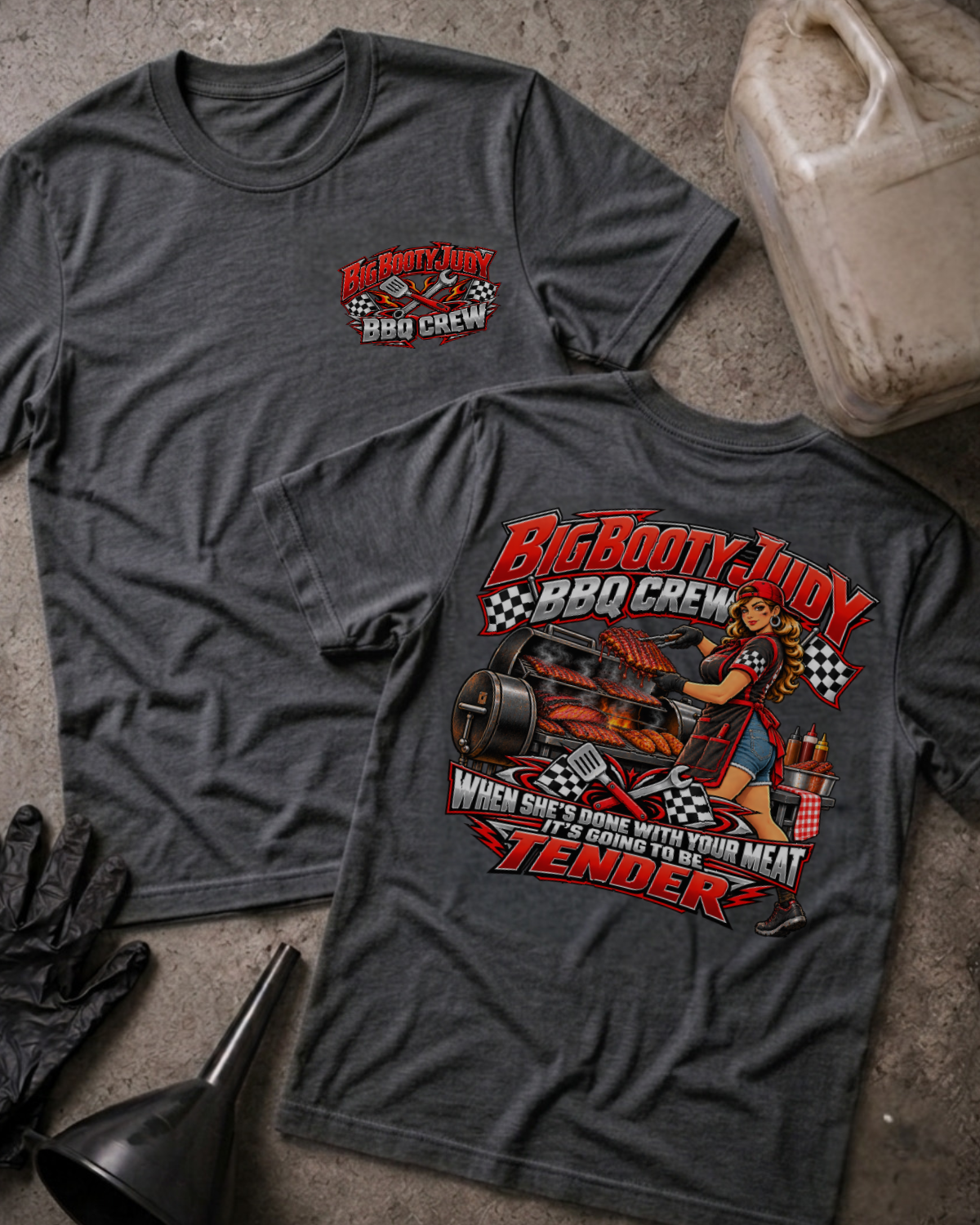 Big Booty Judy BBQ Crew T-shirt in dark grey heather, flat lay showing the front chest logo and the back graphic reading Big Booty Judy BBQ Crew, When She's Done With Your Meat It's Going to Be Tender, styled with work gloves, a funnel, and a fluid jug.