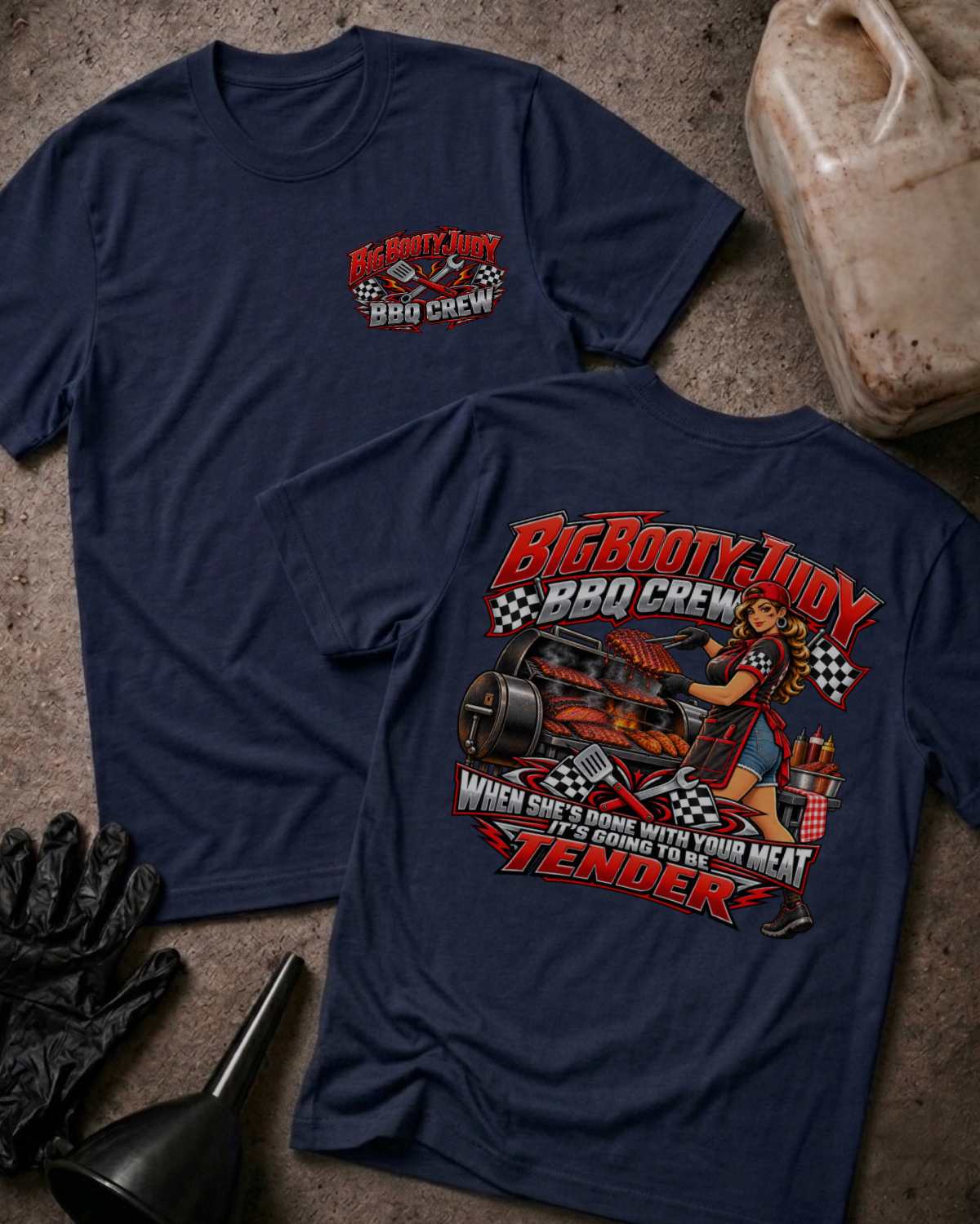 Big Booty Judy BBQ Crew T-shirt in navy, flat lay showing the front chest logo and the back graphic reading Big Booty Judy BBQ Crew, When She's Done With Your Meat It's Going to Be Tender, styled with work gloves, a funnel, and a fluid jug.