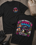 Two black graphic t-shirts displayed flat on a concrete surface, with a race fuel jug, black gloves, and a funnel as props. The front shirt shows a small colorful chest graphic, and the back shirt features a large graphic of a blue race car with the text 'BIG BOOTY JUDY BRAKES' and 'STOPS YOU DEAD IN YOUR TRACKS'.