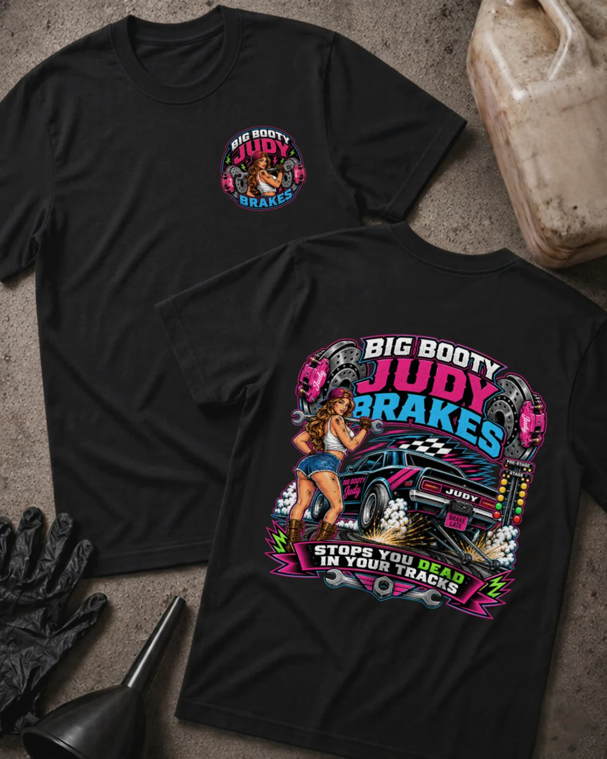 Two black graphic t-shirts displayed flat on a concrete surface, with a race fuel jug, black gloves, and a funnel as props. The front shirt shows a small colorful chest graphic, and the back shirt features a large graphic of a blue race car with the text 'BIG BOOTY JUDY BRAKES' and 'STOPS YOU DEAD IN YOUR TRACKS'.