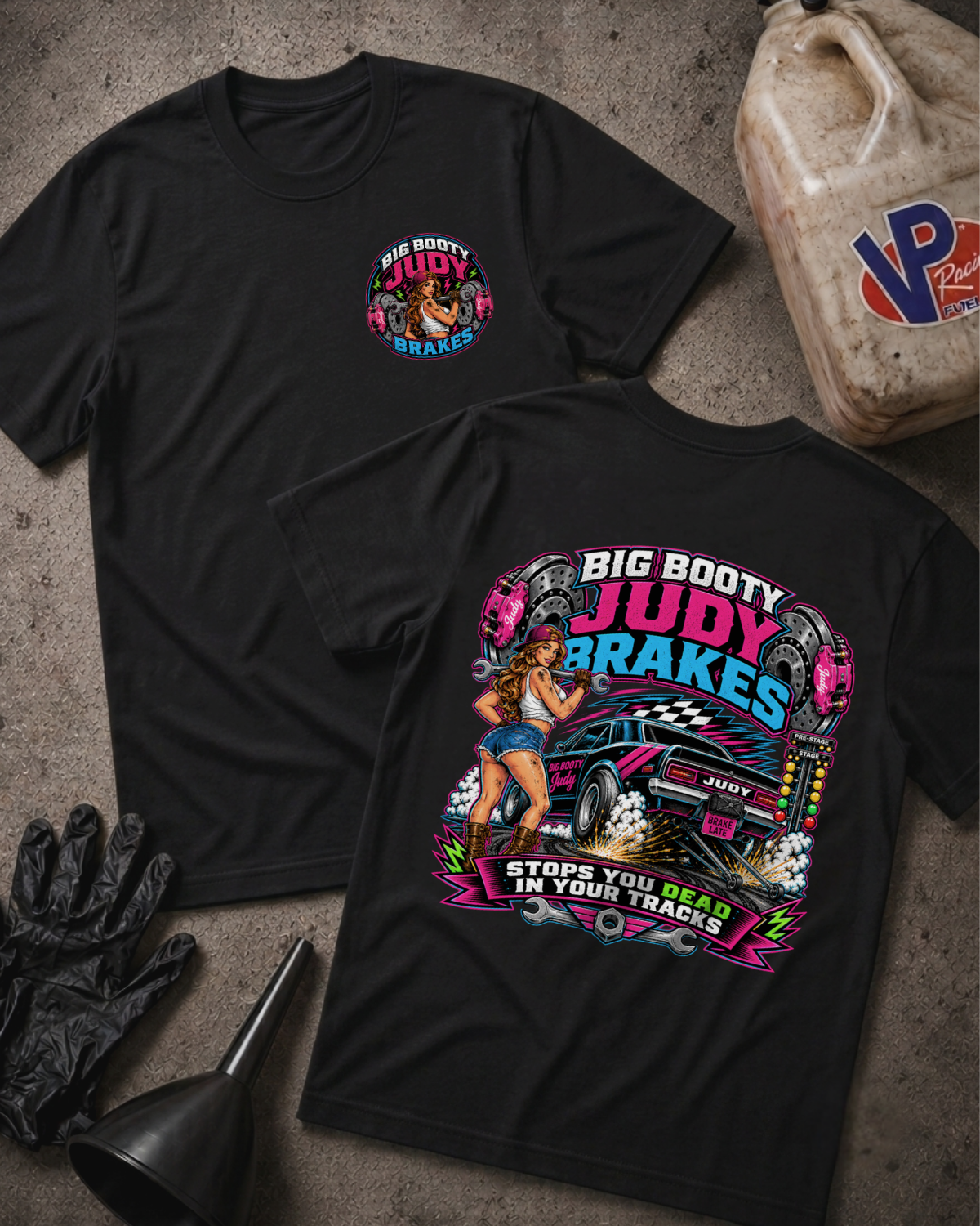 Two black BIG BOOTY JUDY BRAKES graphic T-shirts laid flat on a textured gray surface, with racing-themed printed artwork and text. A black glove, funnel, and a plastic fuel can are placed around them as styled props.