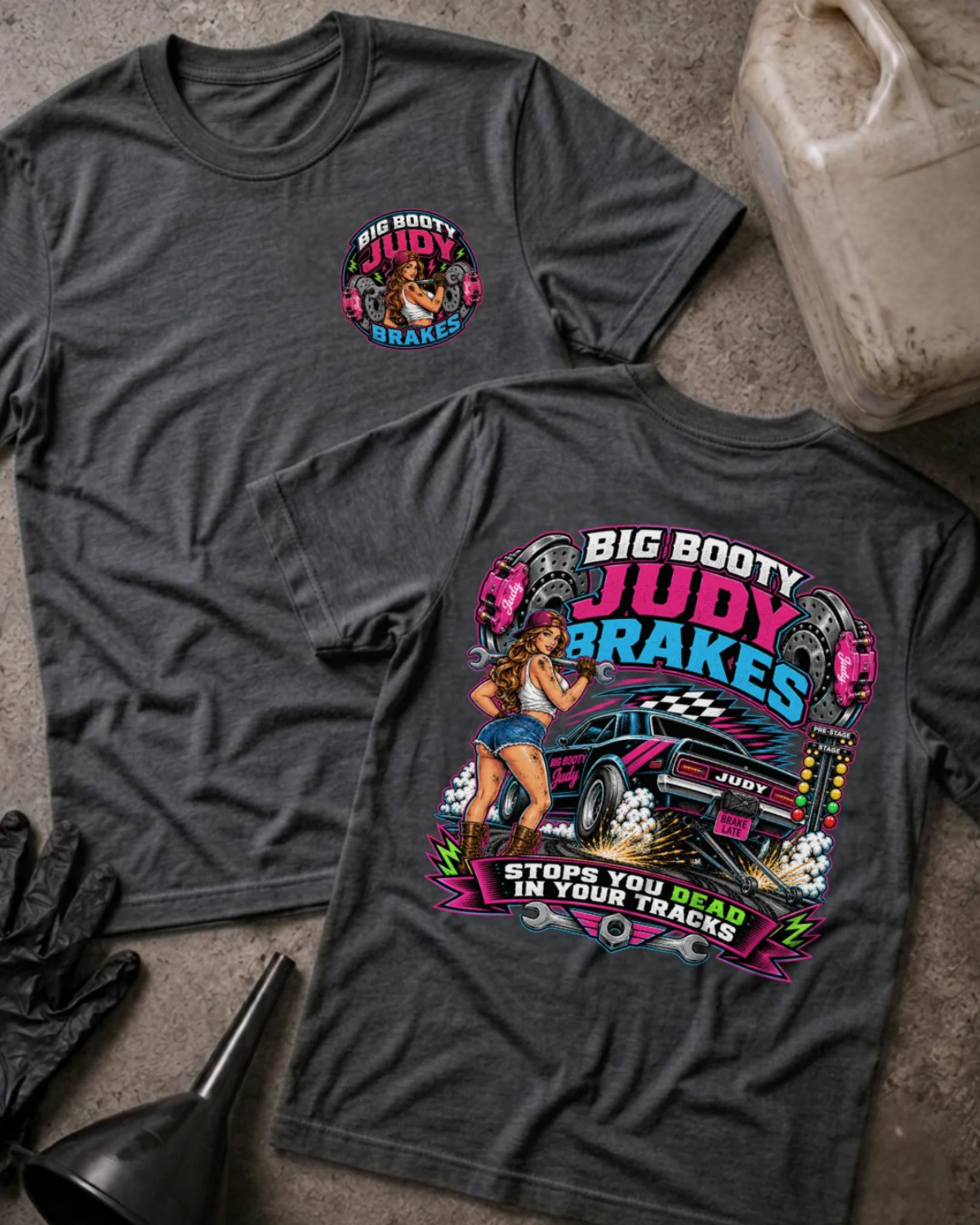 Two dark heather gray graphic T-shirts laid flat on a concrete-like surface, showing front and back designs. The front has a small circular chest graphic reading 'Big Booty Judy Brakes,' and the back has a large colorful pin-up car graphic with the same text.