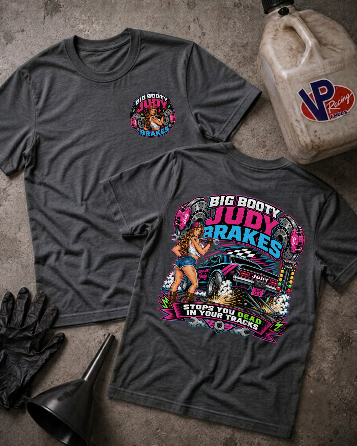 Two dark heather gray graphic t-shirts displayed flat on a concrete surface, with a race fuel jug, black gloves, and a funnel as props. The front shirt shows a small colorful chest graphic, and the back shirt features a large graphic of a blue race car with the text 'BIG BOOTY JUDY BRAKES' and 'STOPS YOU DEAD IN YOUR TRACKS'.