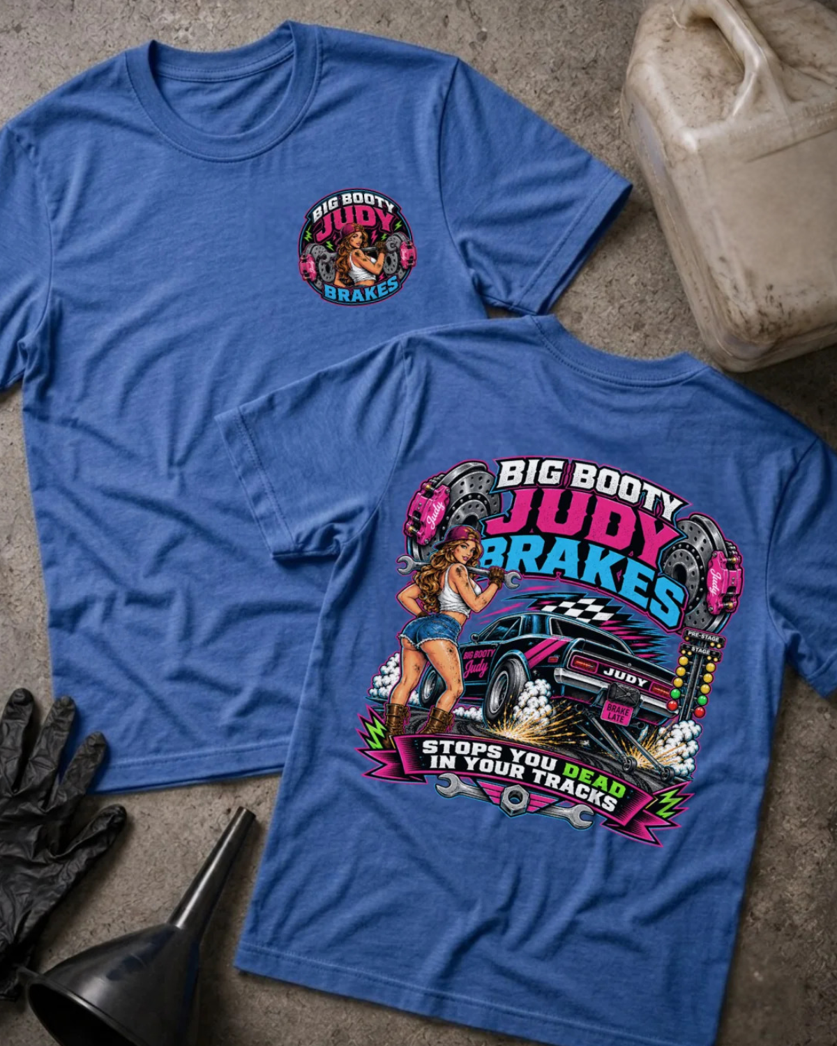 Two blue graphic T-shirts laid flat on a textured surface, showing the front and back designs. The front has a small circular chest graphic, and the back has a large colorful graphic with the text 'BIG BOOTY JUDY BRAKES' and a pin-up style illustration.